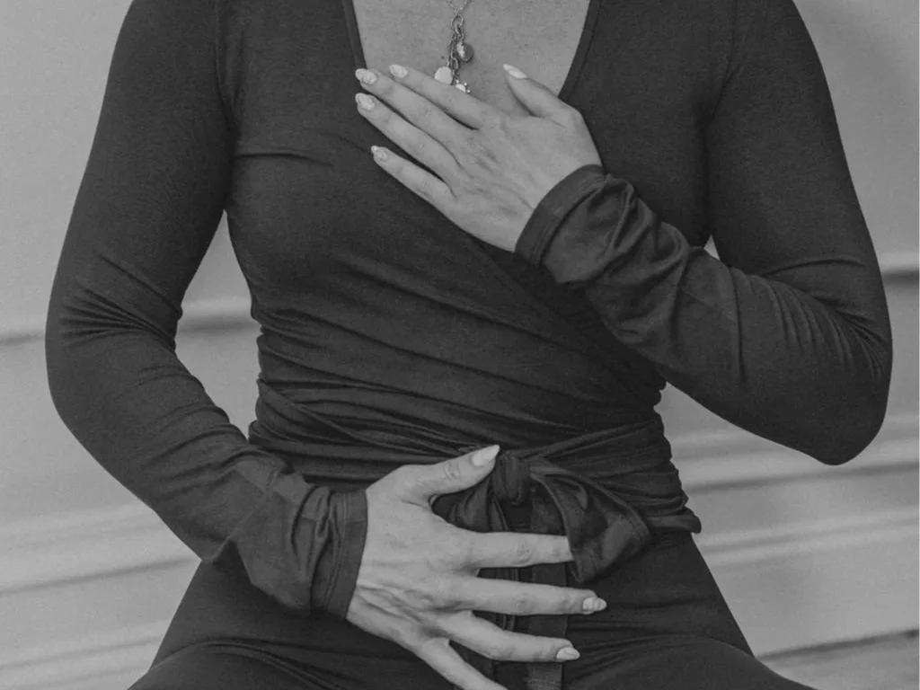 Close-up of a woman with one hand on her chest and the other on her stomach, both hands resting over her midsection, wearing a dark long-sleeve shirt with a knot at the waist.
