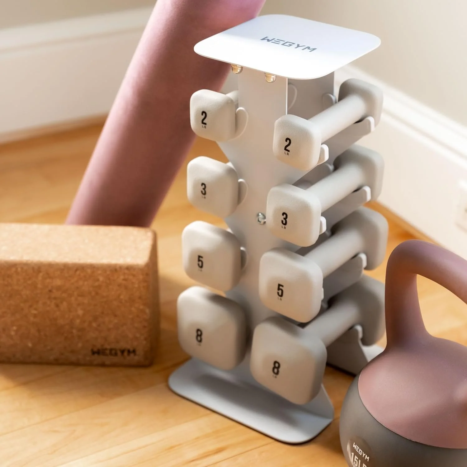 Set of HEGYM dumbbells with various weights stacked on a white rack, a pink yoga ball, and a cork yoga block on a wooden floor.