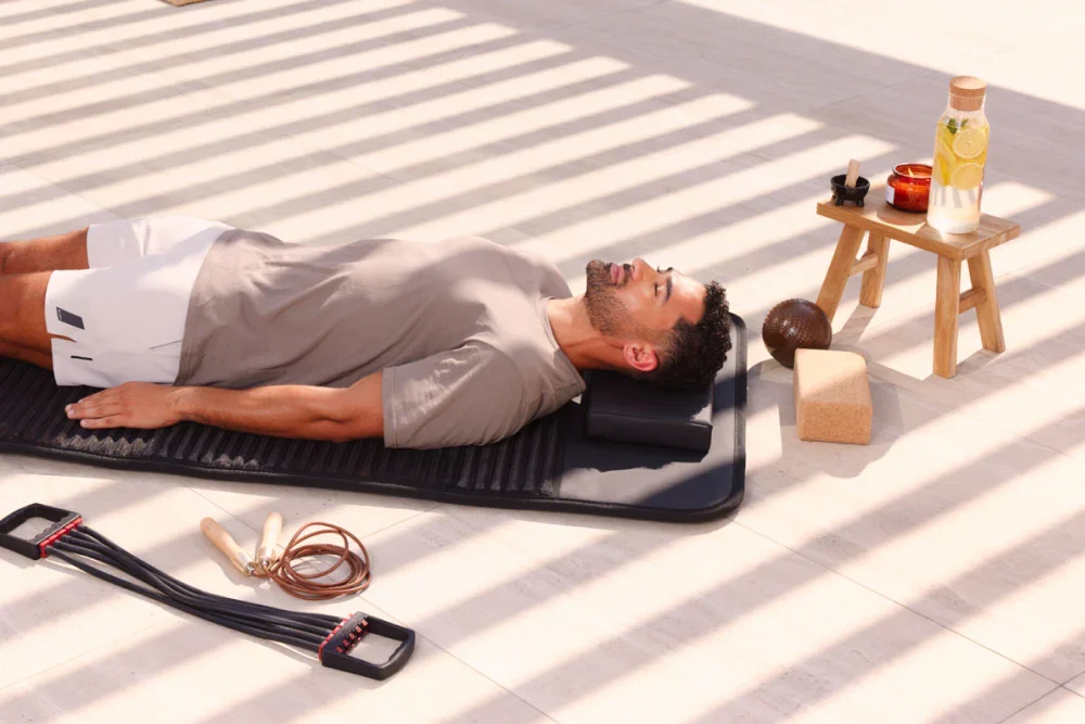 A man lying on his back on a yoga mat, relaxing with eyes closed. There are yoga props and accessories nearby, including a wooden stool with a drink and candles, a massage ball, a cork, a sponge, and resistance bands.
