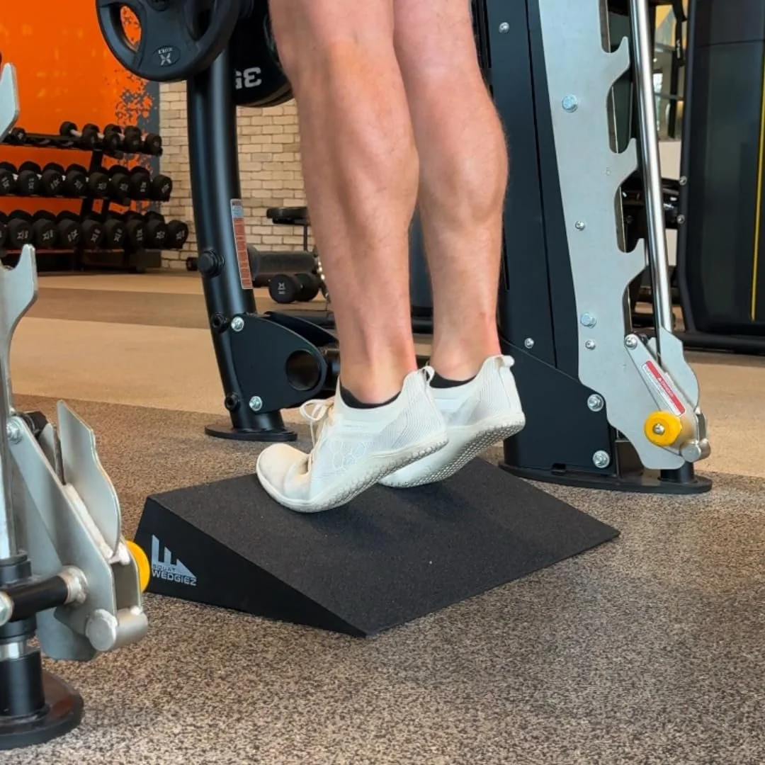 Close-up of a person performing calf raises on a slanted wedge at the gym, with gym equipment and dumbbells in the background.