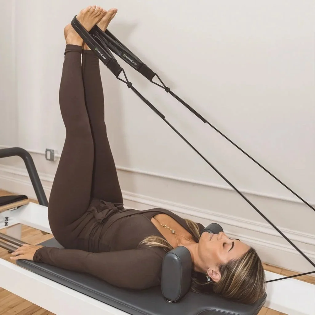 Woman lying on her back on a Pilates reformer, using resistance bands for exercise, with her legs elevated and her head resting on padded supports.