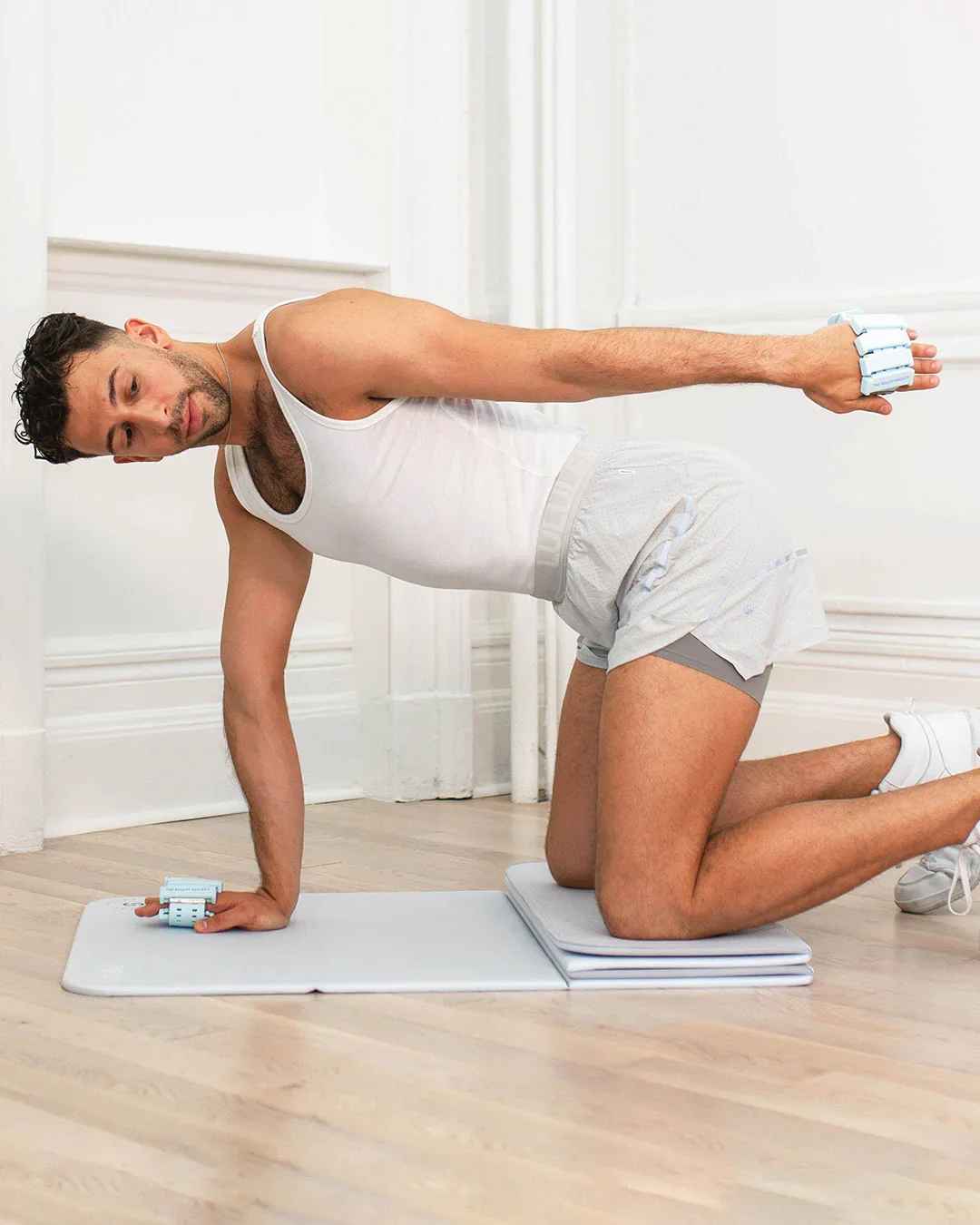 Man performing a yoga pose on a mat, wearing a white tank top and shorts, with ankle weights.
