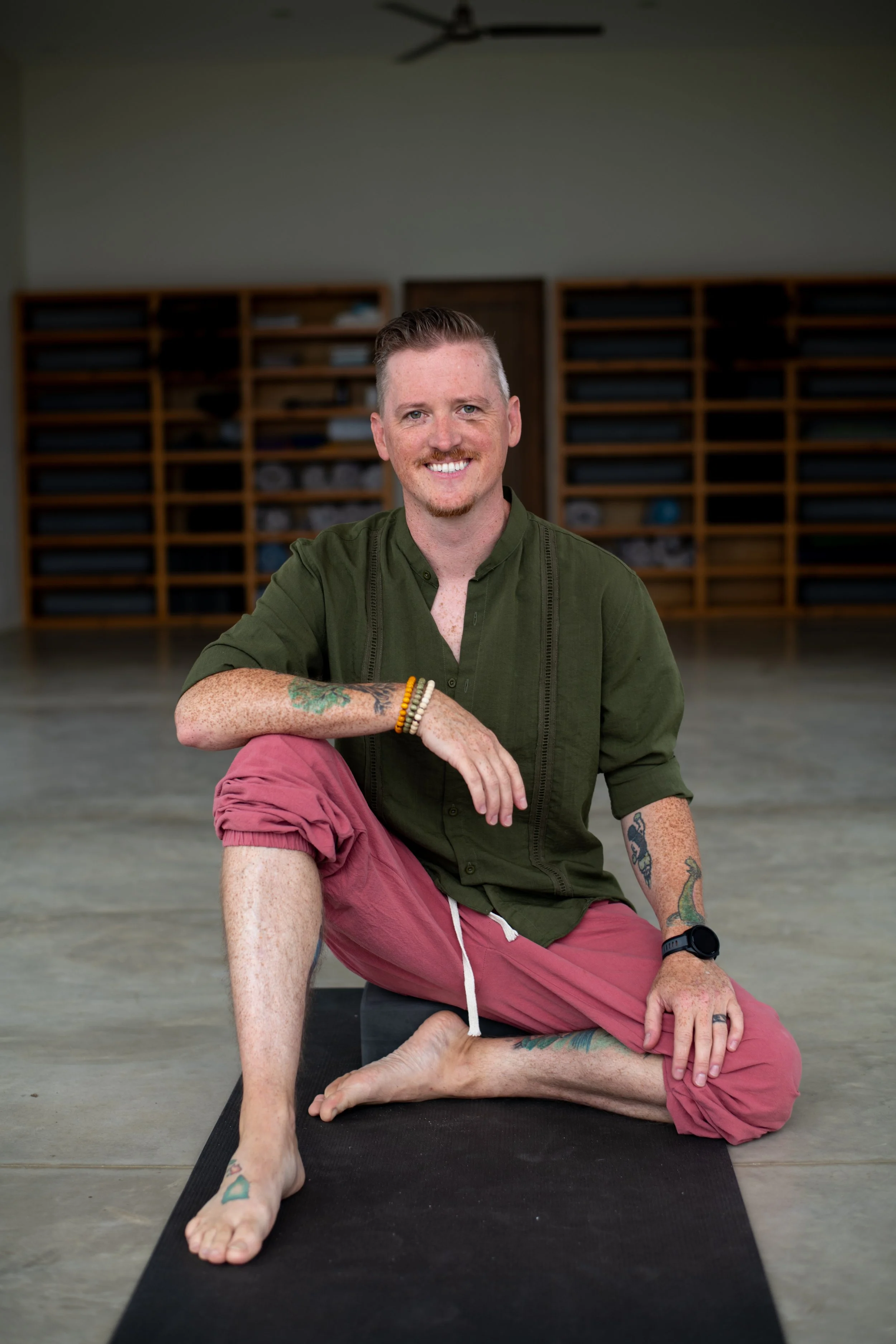 A man sitting on a yoga mat in a yoga studio, smiling at the camera, wearing a green shirt and pink pants, with tattoos on his arms and legs.