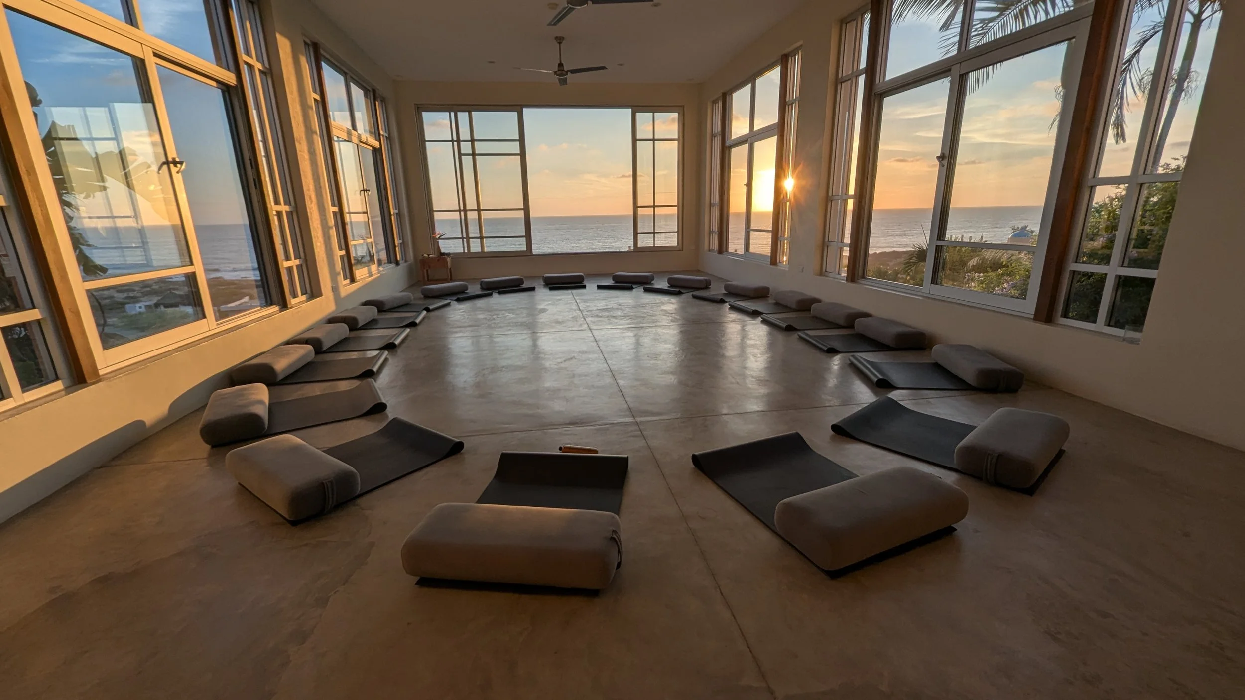 Sunset view through large windows in a yoga studio with black mats and gray bolsters arranged in a circle.