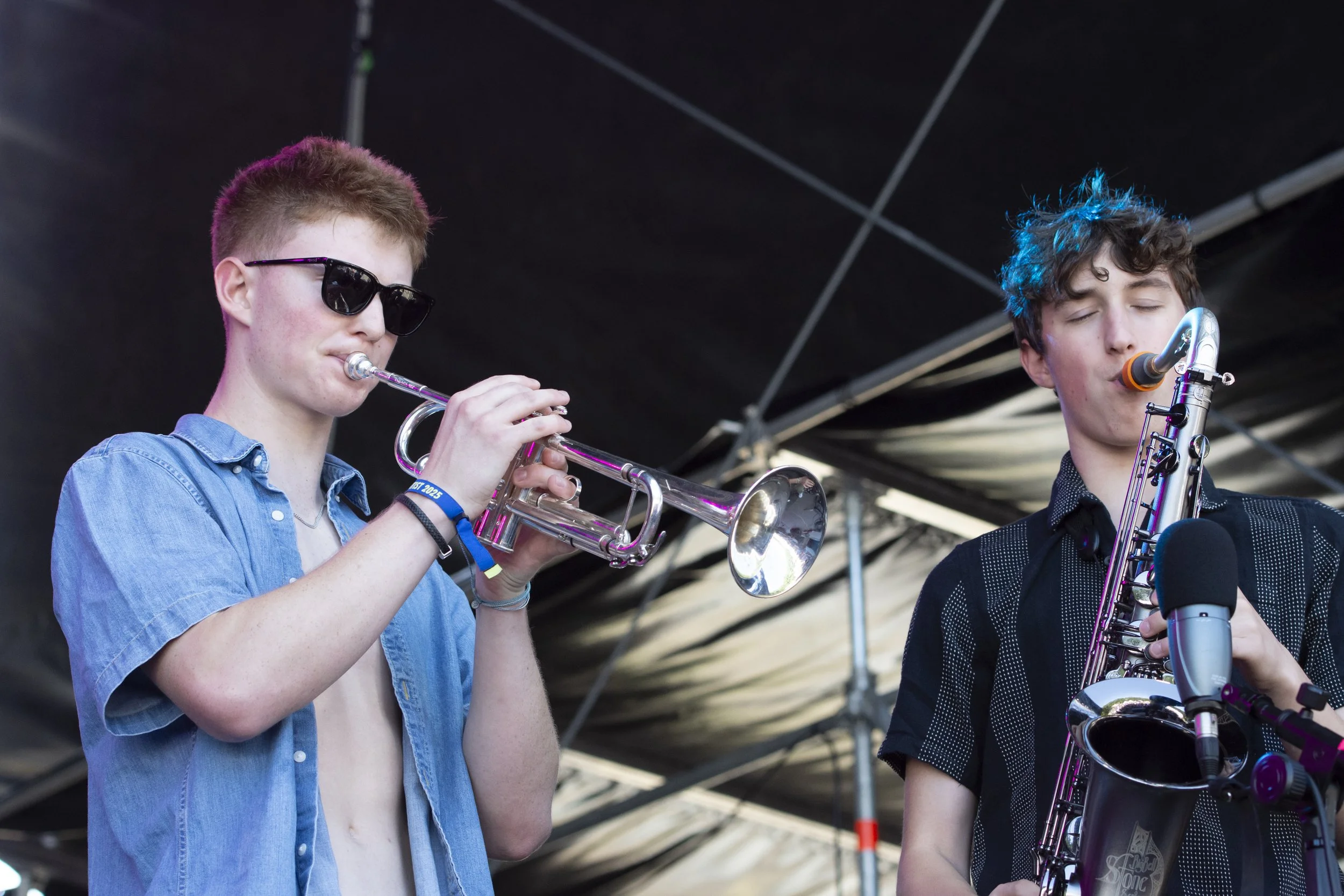 Two young male musicians performing on stage, one playing a trumpet and the other playing a saxophone, under a dark tent roof.