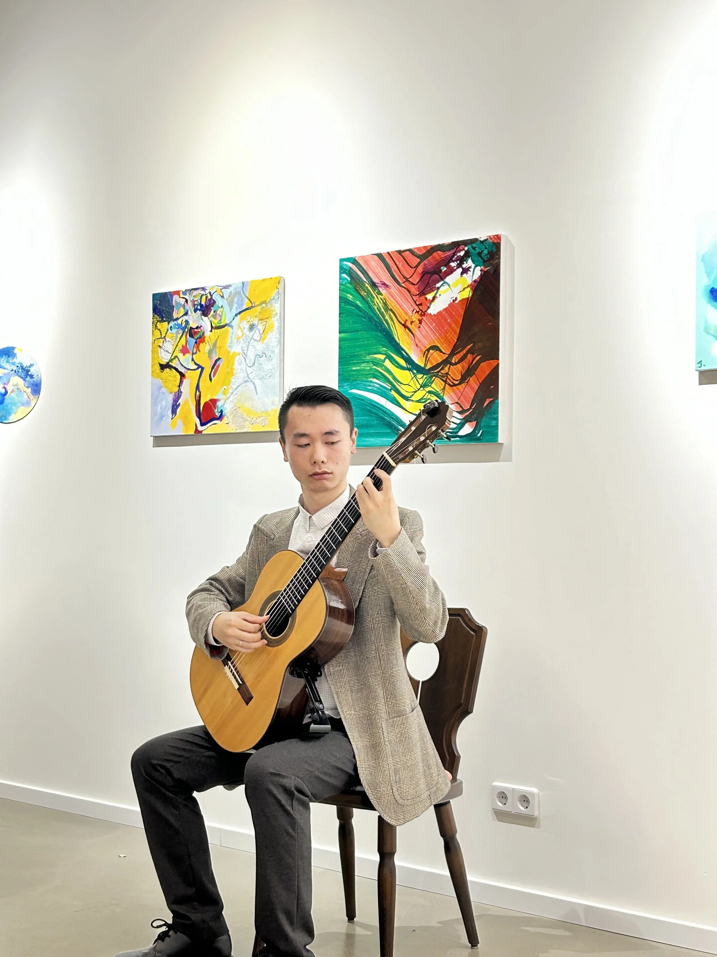A young man playing an acoustic classical guitar while sitting on a wooden chair in an art gallery with colorful paintings on the wall.