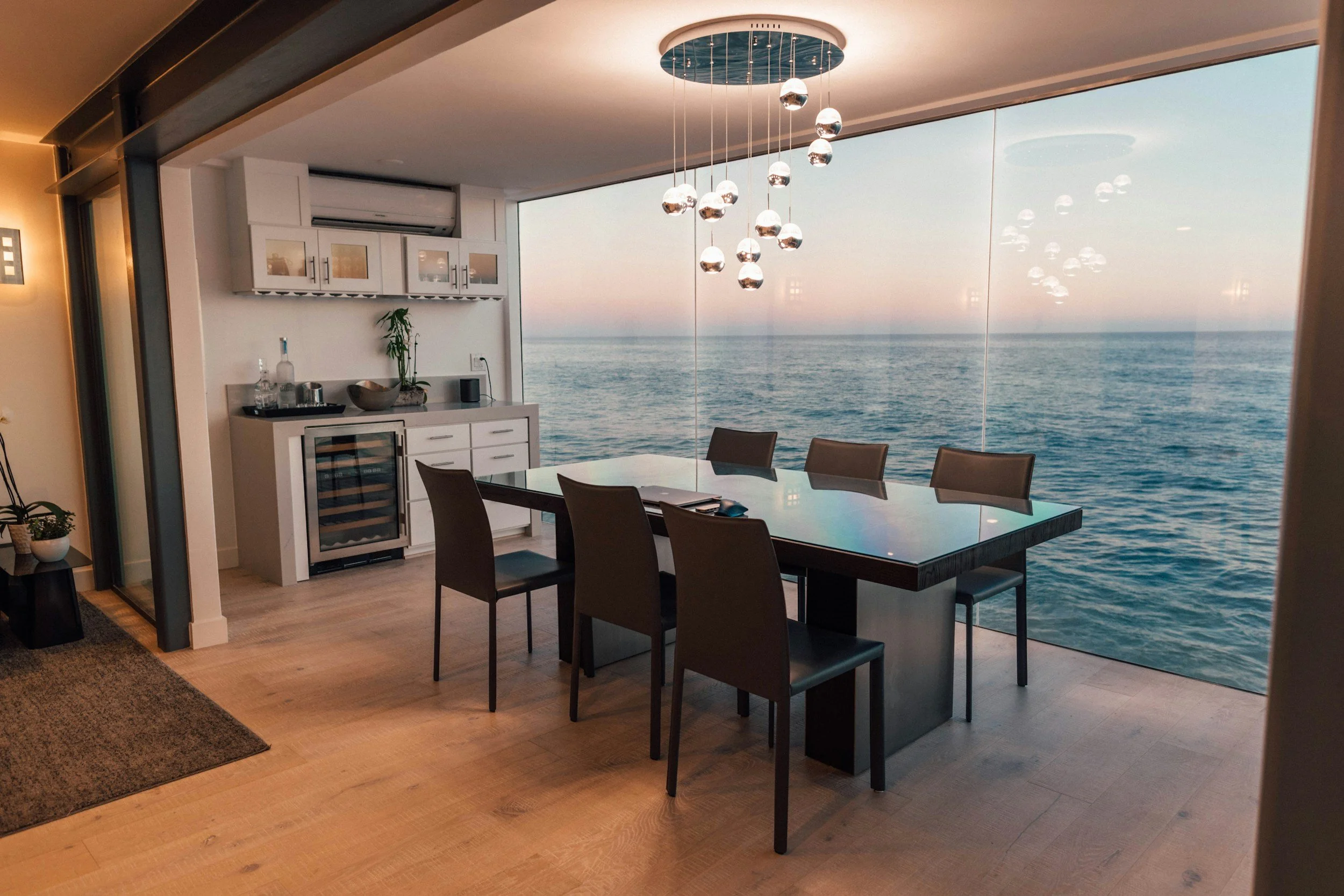 Dining room with a large window overlooking the ocean during sunset, contemporary chandelier hanging above a dark wooden dining table with six chairs, and a small kitchenette with a wine cooler and decorative items in the background.