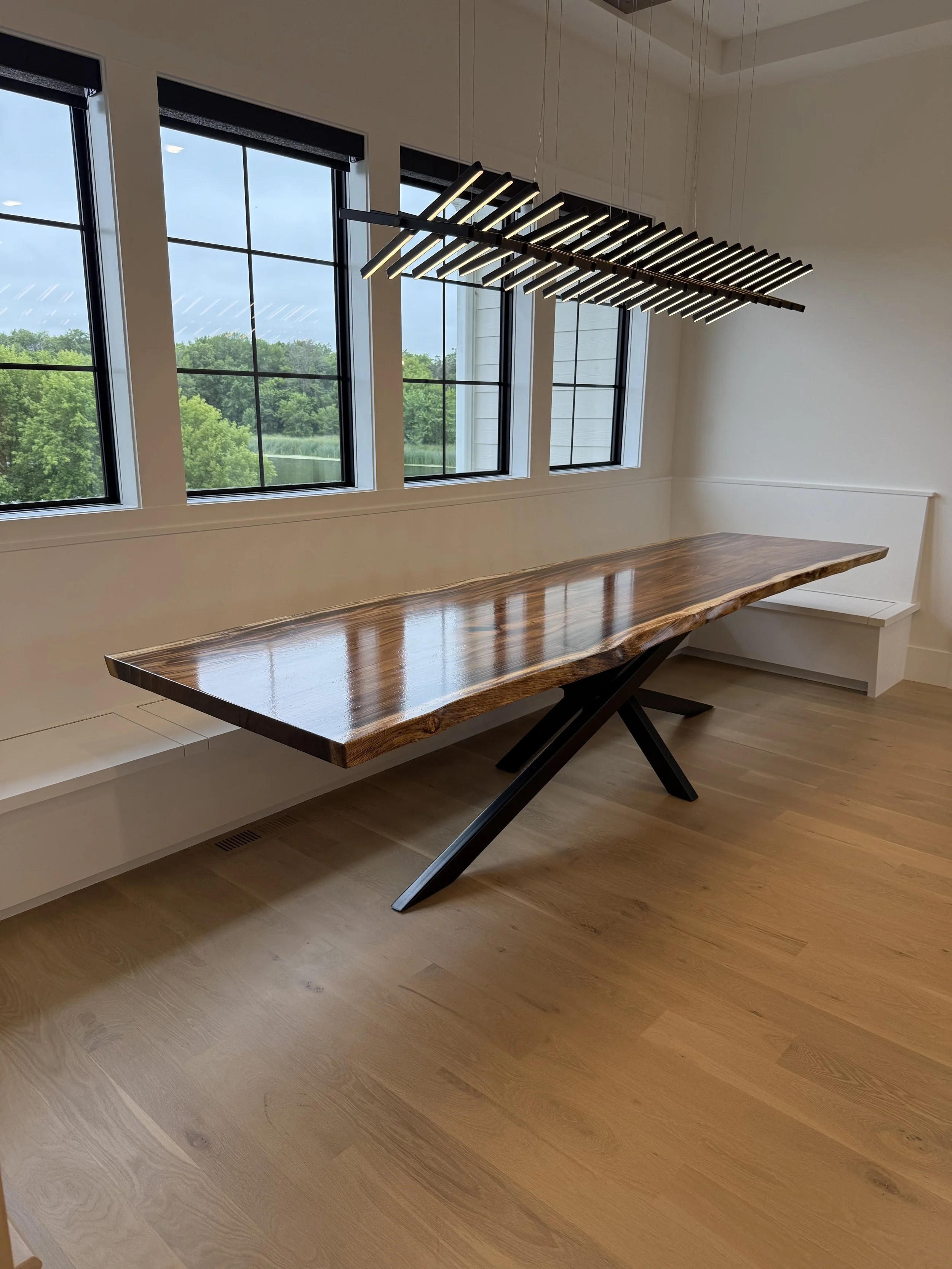 A modern dining area with a live edge wooden table, black metal legs, three large windows with black blinds, a sleek black hanging chandelier, and a white built-in bench along the wall. Outside scenery of trees is visible through the windows.