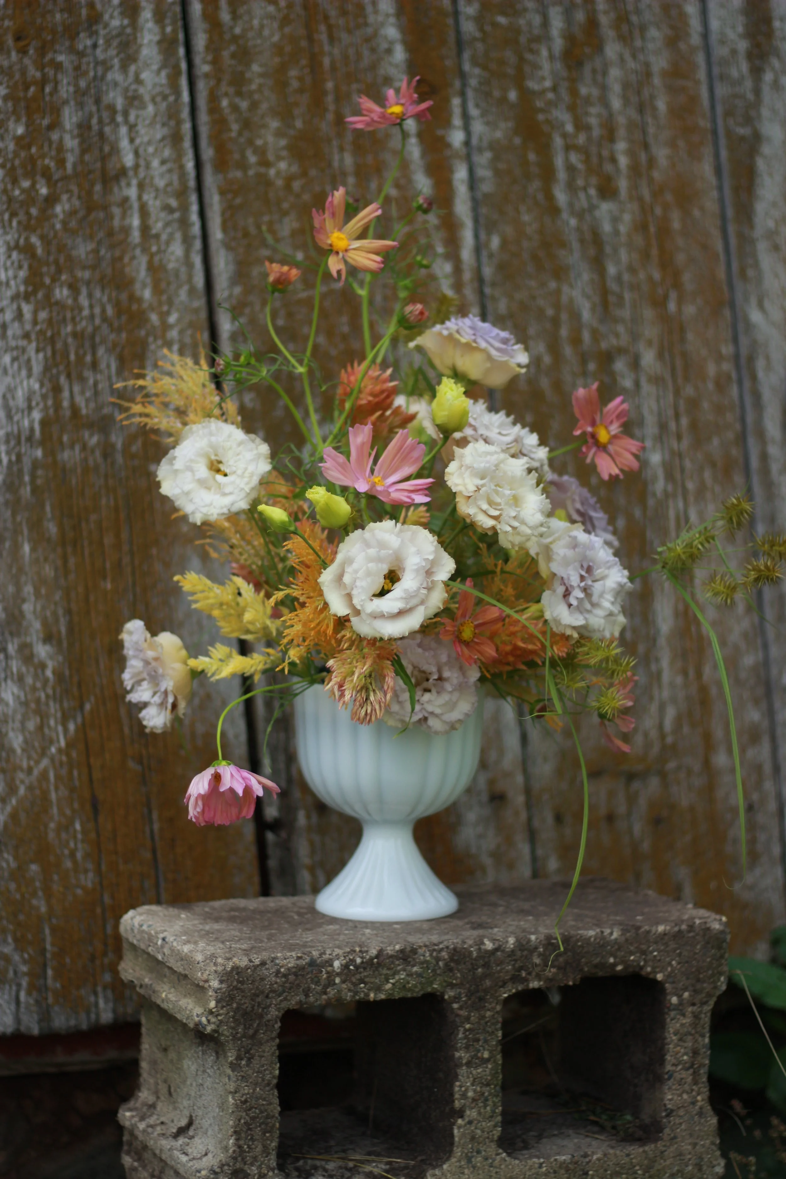 A bouqet of flowers in a white goblet vase sitting on a cinder block in front of a stressed-wood paneled background