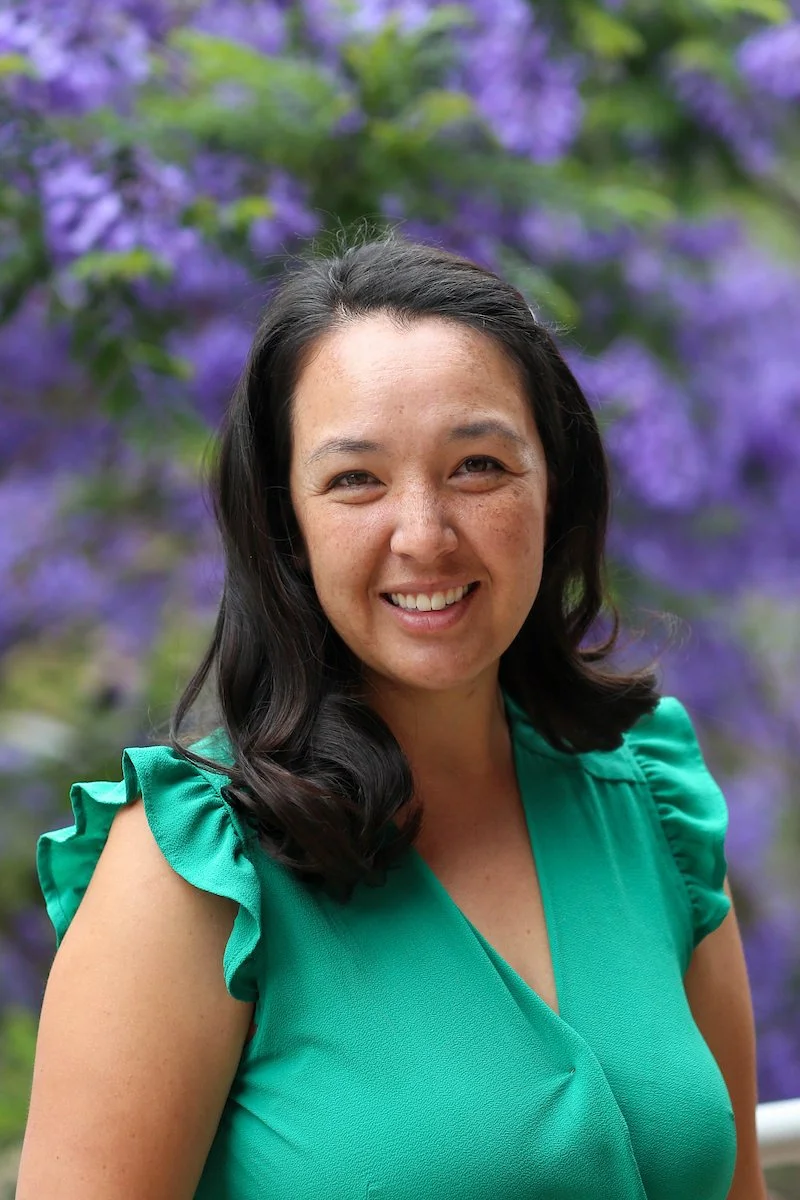 A woman with dark, wavy hair smiling outdoors in front of purple flowering bushes, wearing a teal top with ruffled sleeves.
