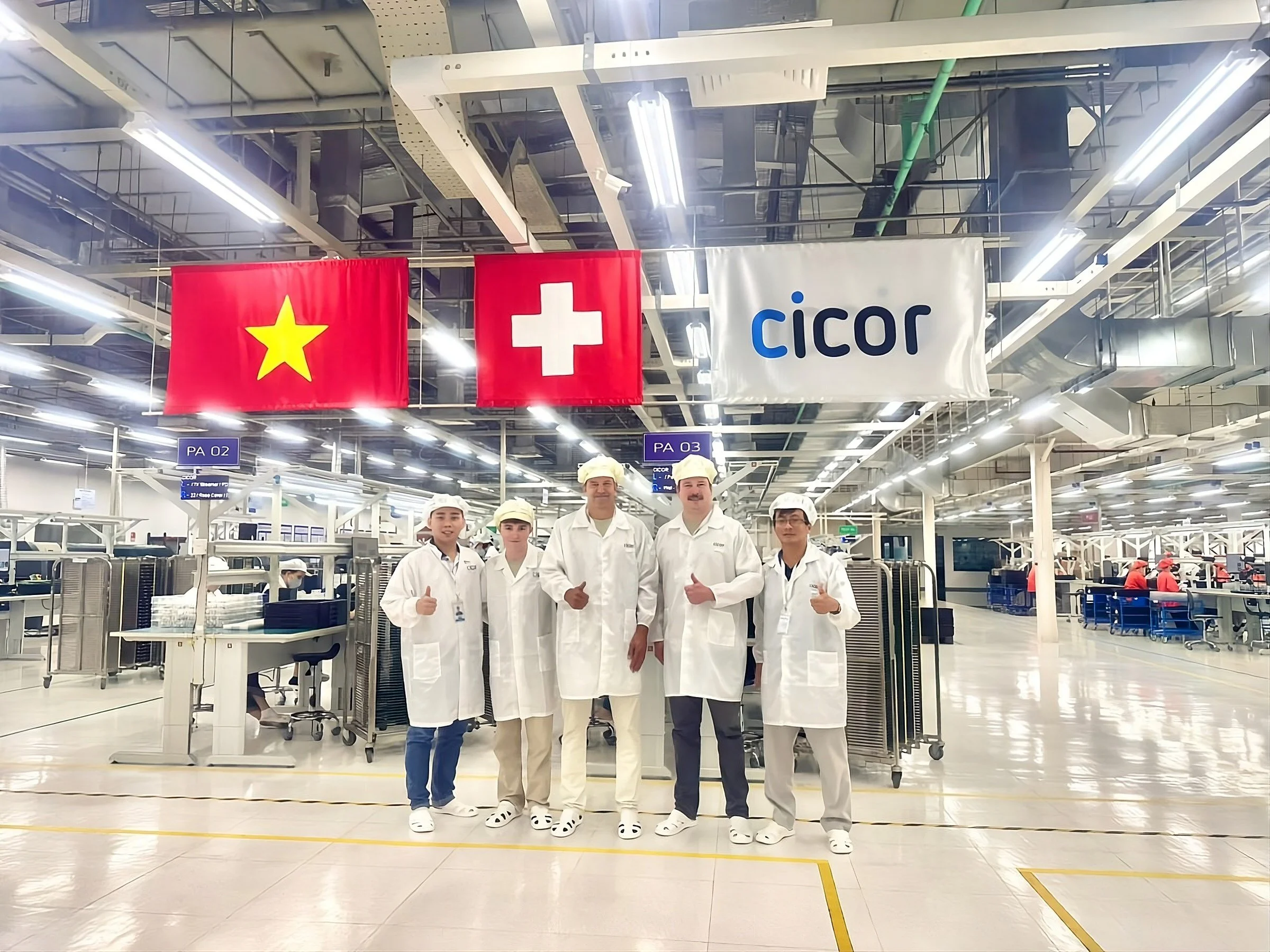 Group of five workers wearing white protective clothing, hairnets, and safety shoes, standing in a manufacturing or food processing facility with flags of Vietnam, Switzerland, and a company banner that reads 'Cicor' hanging above.