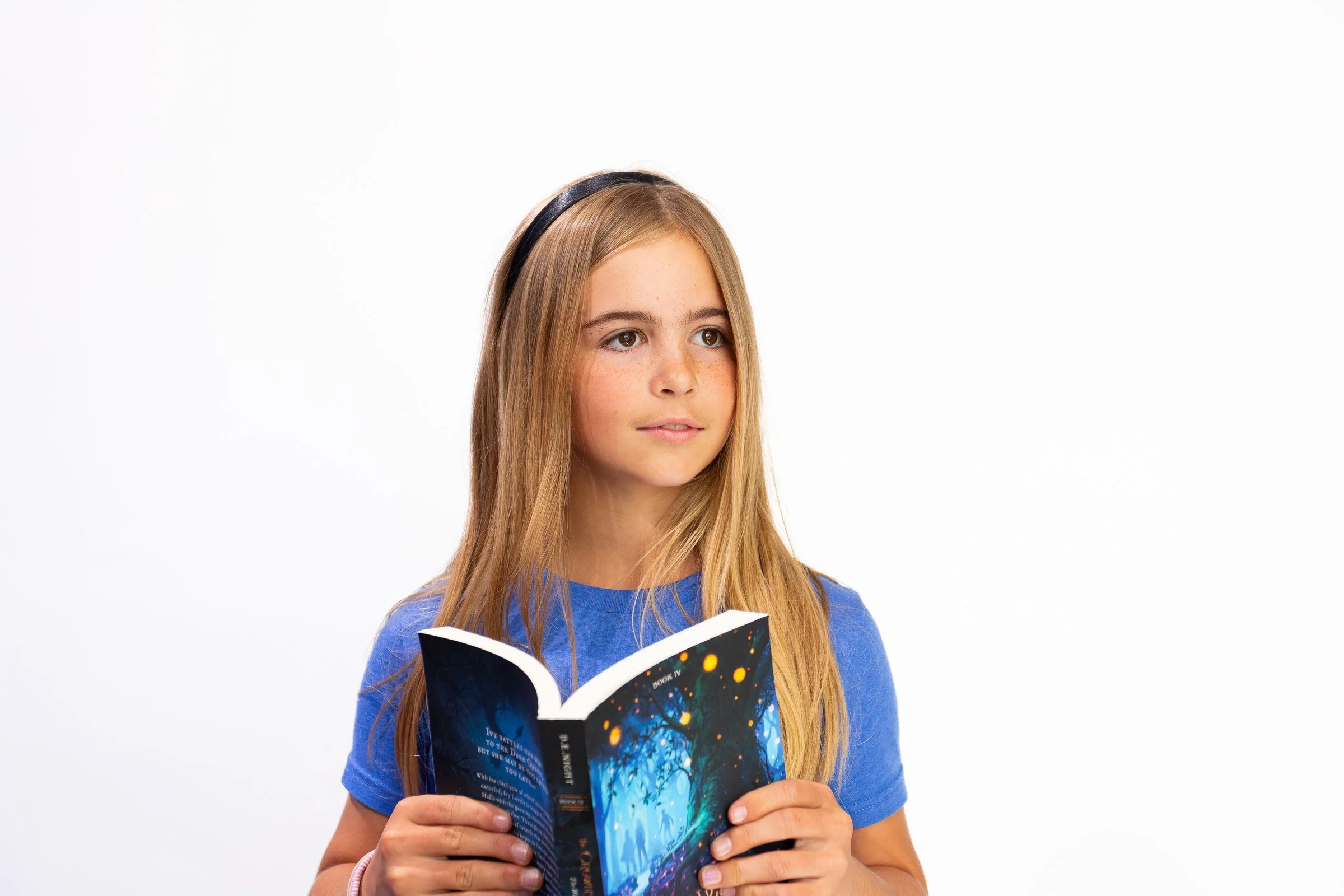 A young girl with long light brown hair, blue shirt, and headband reads a colorful book with a galaxy and trees cover against a plain white background.