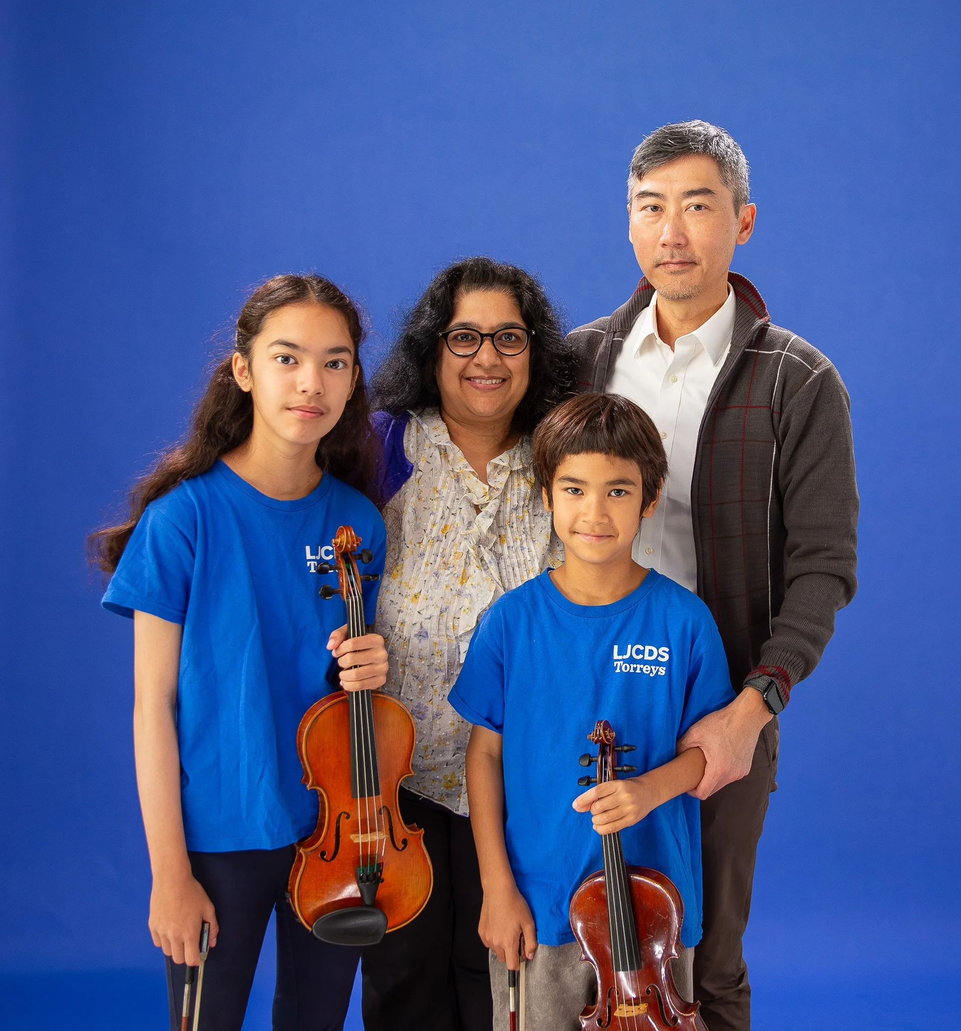 Group portrait of five diverse individuals, including two children holding violins, posing against a blue background.