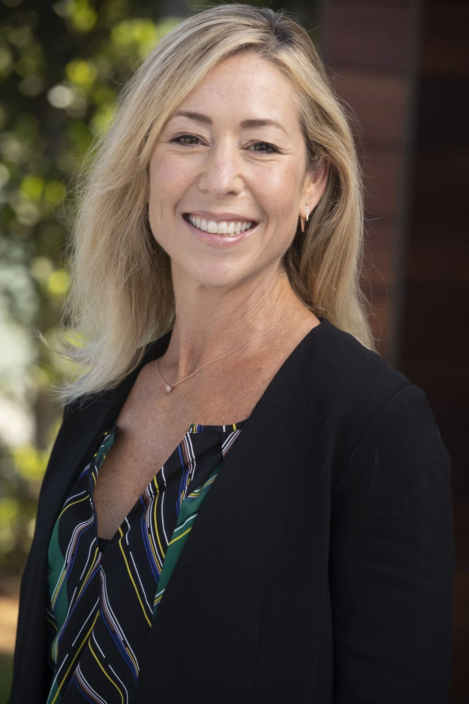 A smiling blonde woman in a black blazer and colorful striped blouse, standing outdoors with greenery in the background.