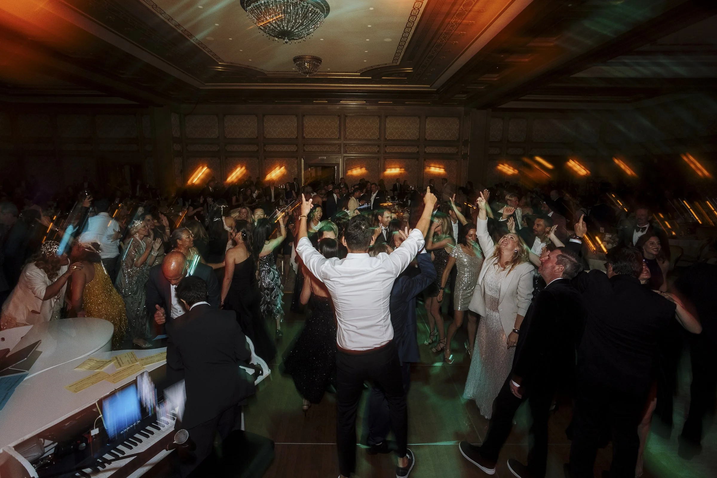 A crowded dance floor with people in formal attire dancing at a celebration or event in an elegant ballroom with chandeliers and warm lighting.