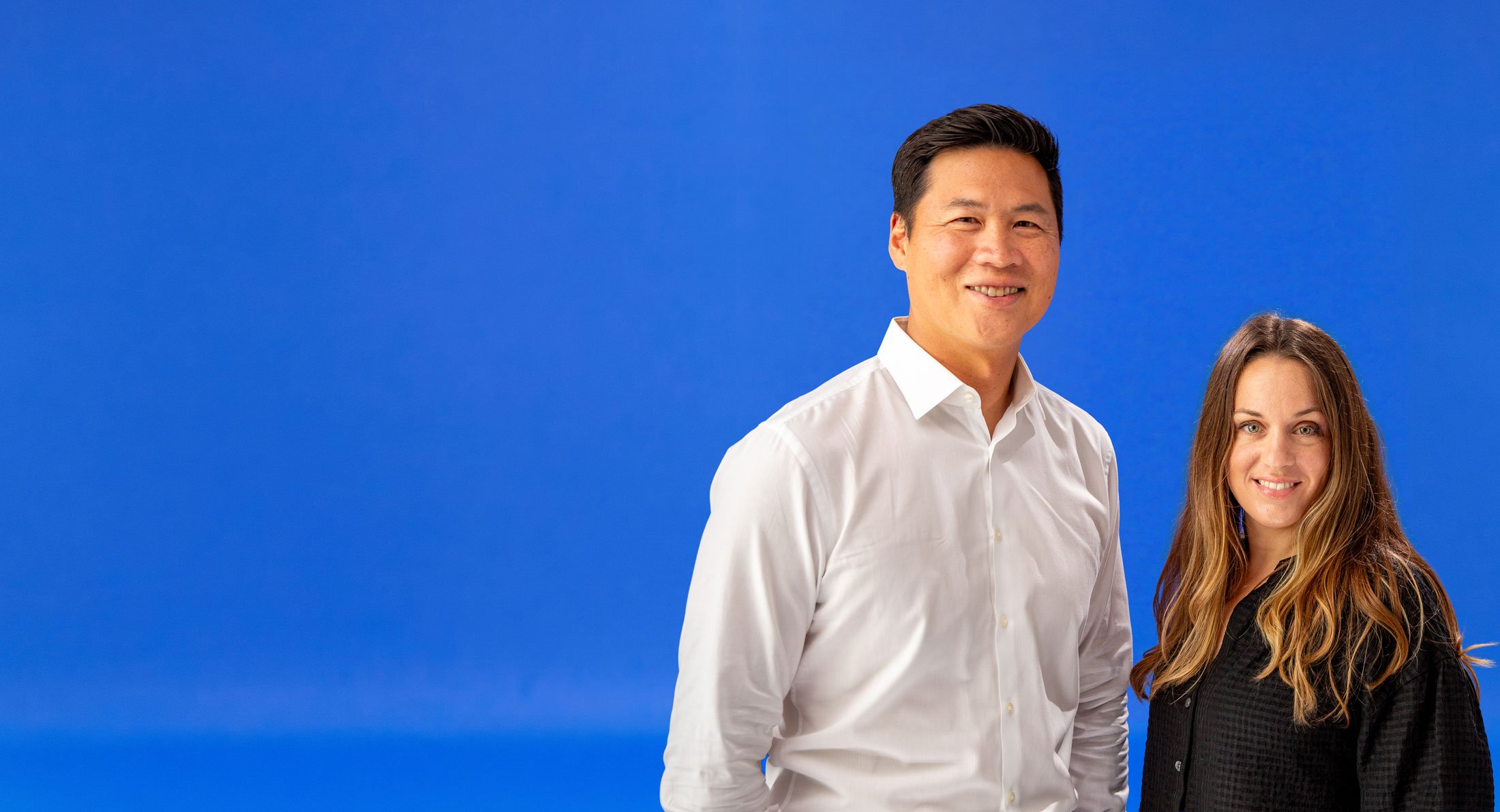 A man in a white shirt standing next to a woman with long brown hair in a black shirt, both smiling, against a solid blue background.
