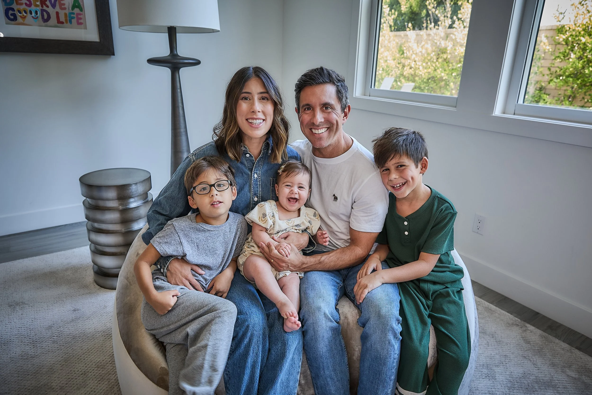 Family of five sitting on a beige sofa in a bright living room with large windows showing trees outside, smiling at the camera.