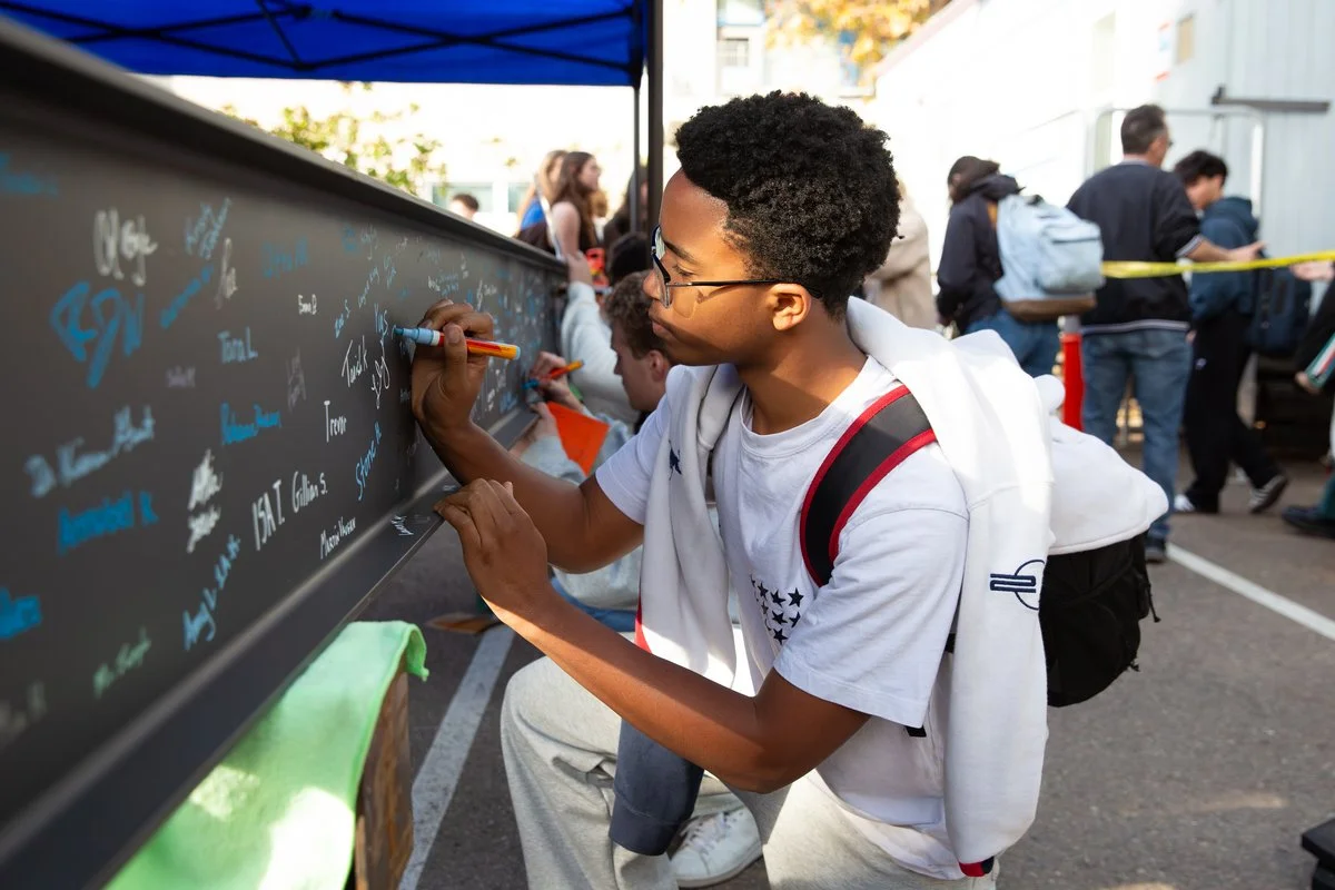 A young boy with glasses writing on a black chalkboard at an outdoor event.