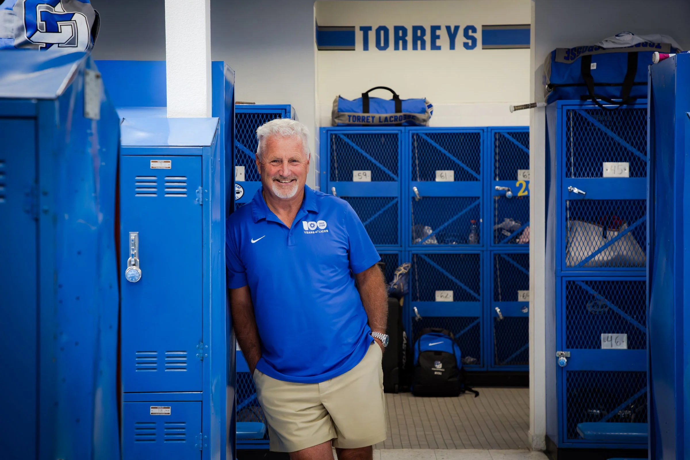 A smiling older man with white hair and a beard, dressed in a blue polo shirt and beige shorts, standing in front of blue lockers in a locker room. The lockers have bags and personal items on top, with signs indicating locker numbers. The background includes a sign with the word 'TORREYS' and sports gear, suggesting a sports or team setting.
