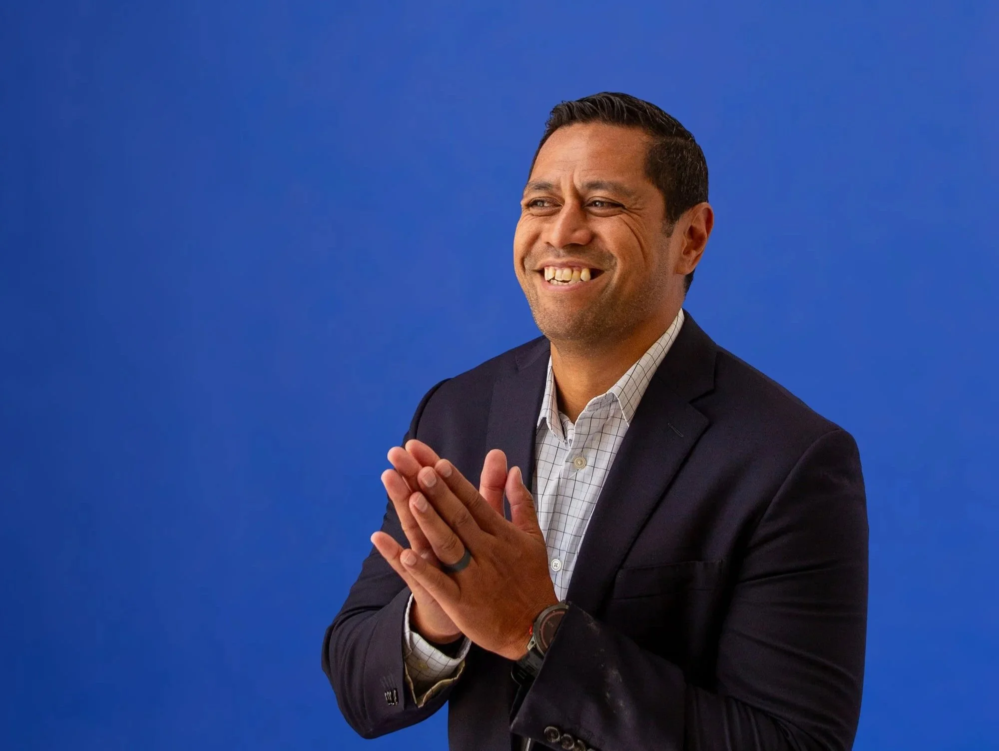 A man smiling, wearing a dark blazer, a light-colored checked shirt, and a watch, standing in front of a blue background.