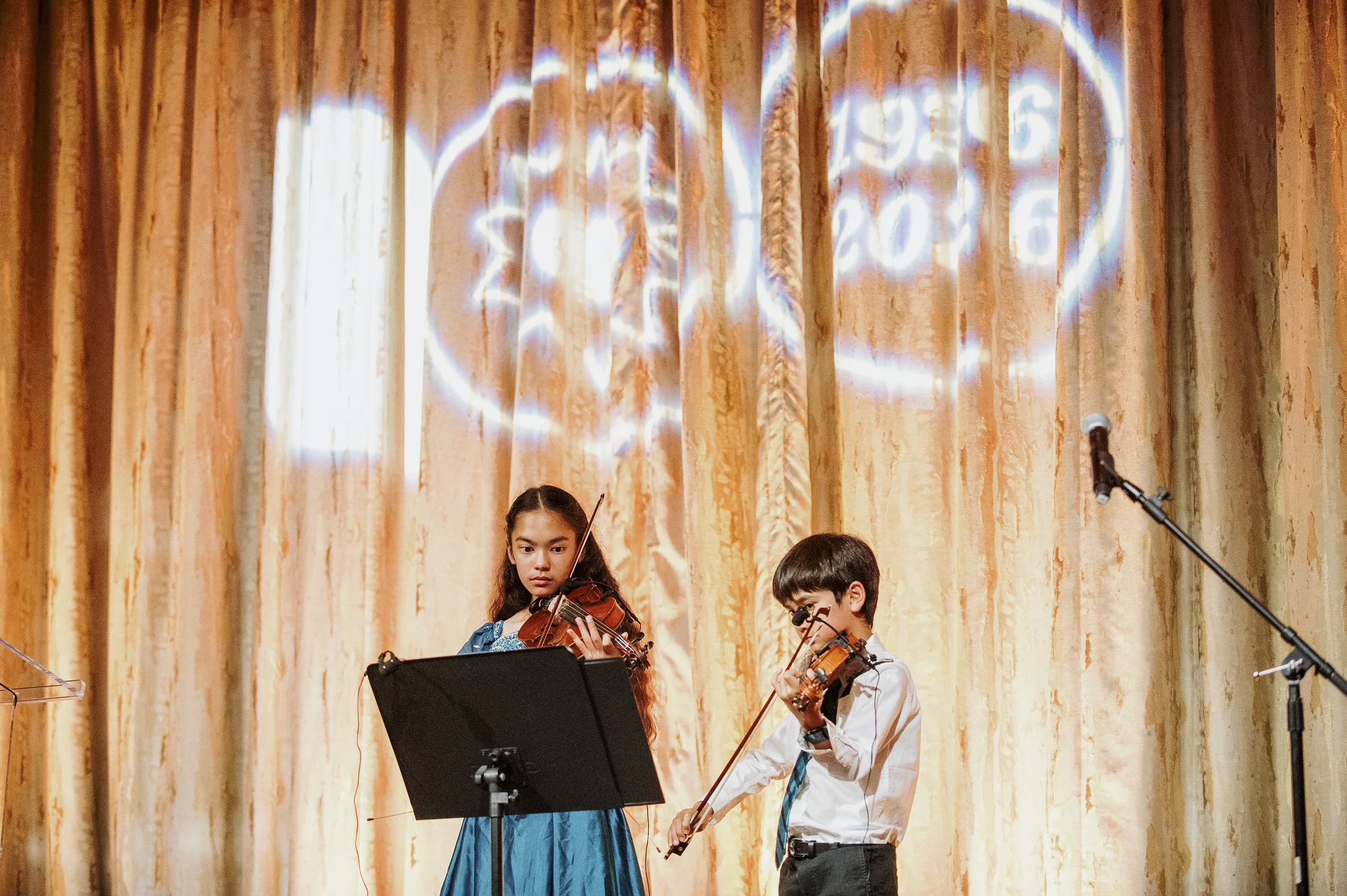Young girl and boy playing violin on stage with yellow curtains in background.