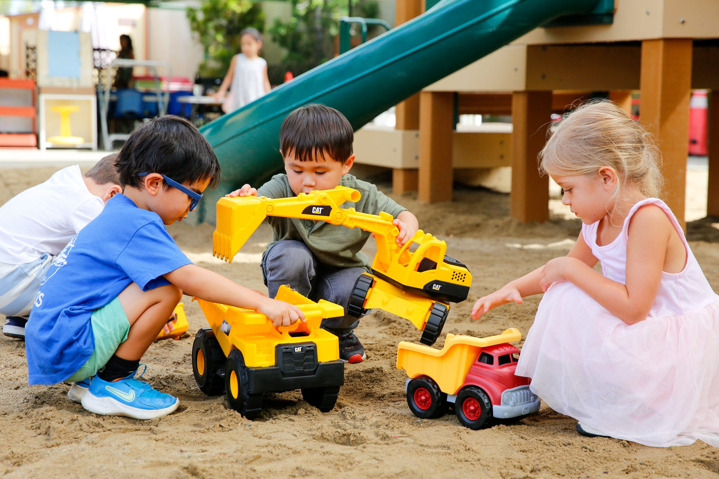 Children playing in sandbox with toy construction vehicles, including a yellow excavator and red dump truck, with a playground slide in background.