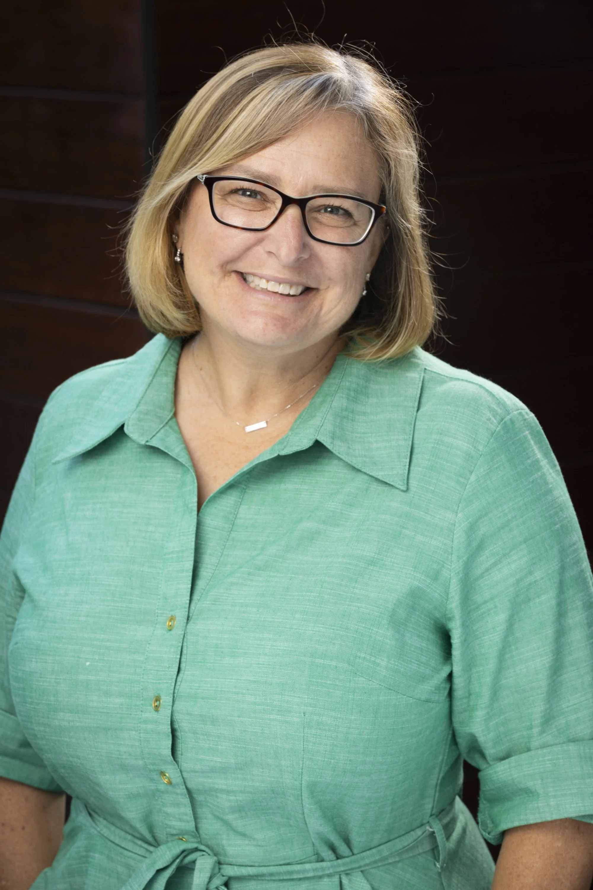 A smiling woman with shoulder-length blonde hair, wearing glasses and a green button-up dress with a tied waist, posing in front of a dark brown wooden background.