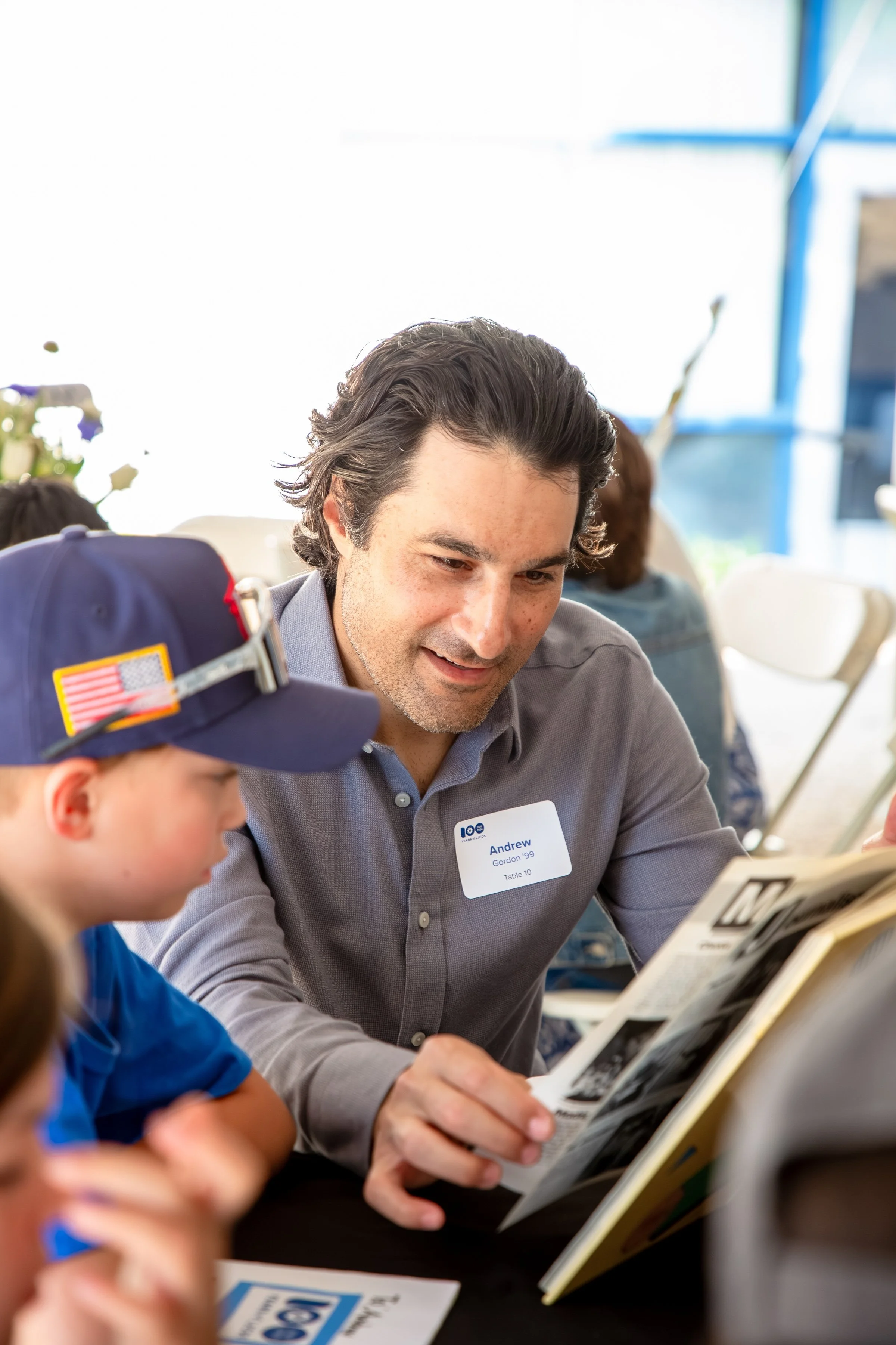 A man with dark hair and a gray shirt is sitting at a table, reading a book to a young boy wearing a blue shirt and a cap with an American flag patch. The man has a name tag that says Andrew Gordon '99, Table 10. The background shows a well-lit room with other people, and some flowers in the distance.