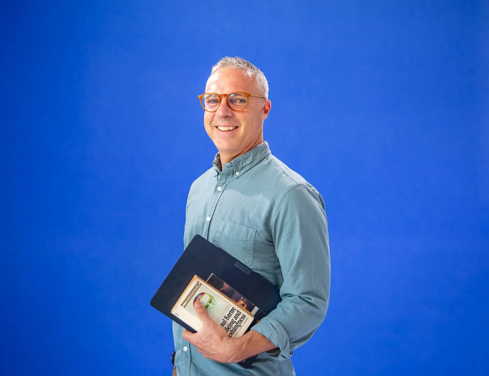 A smiling middle-aged man with glasses and gray hair, wearing a light blue button-up shirt, holding a book and a framed certificate or award, standing against a solid blue background.