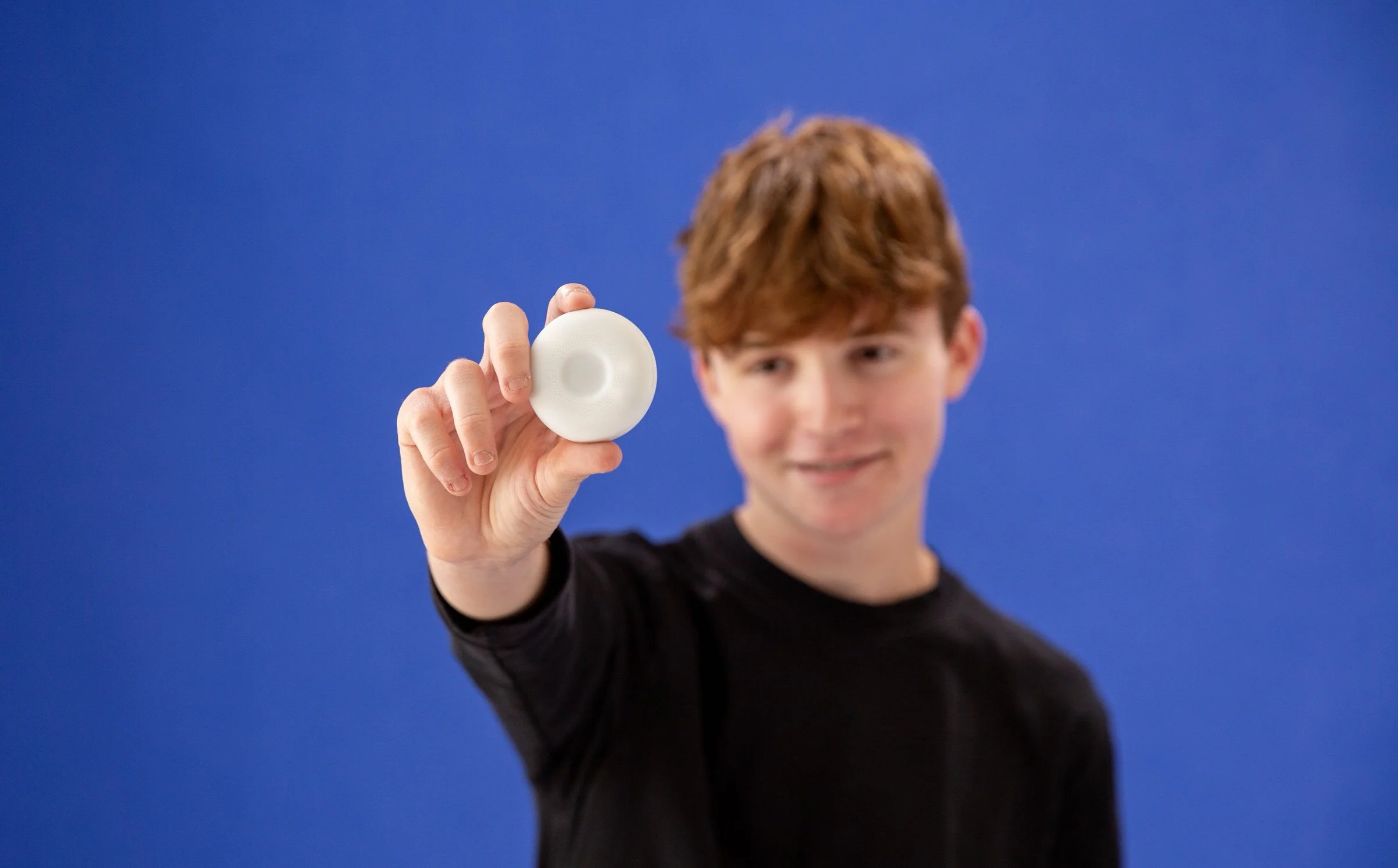 Young man with auburn hair smiling and holding a small white object in front of a solid blue background.