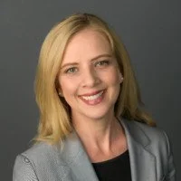 Headshot of a smiling woman with shoulder-length blonde hair, wearing a light gray blazer and black top against a gray background.