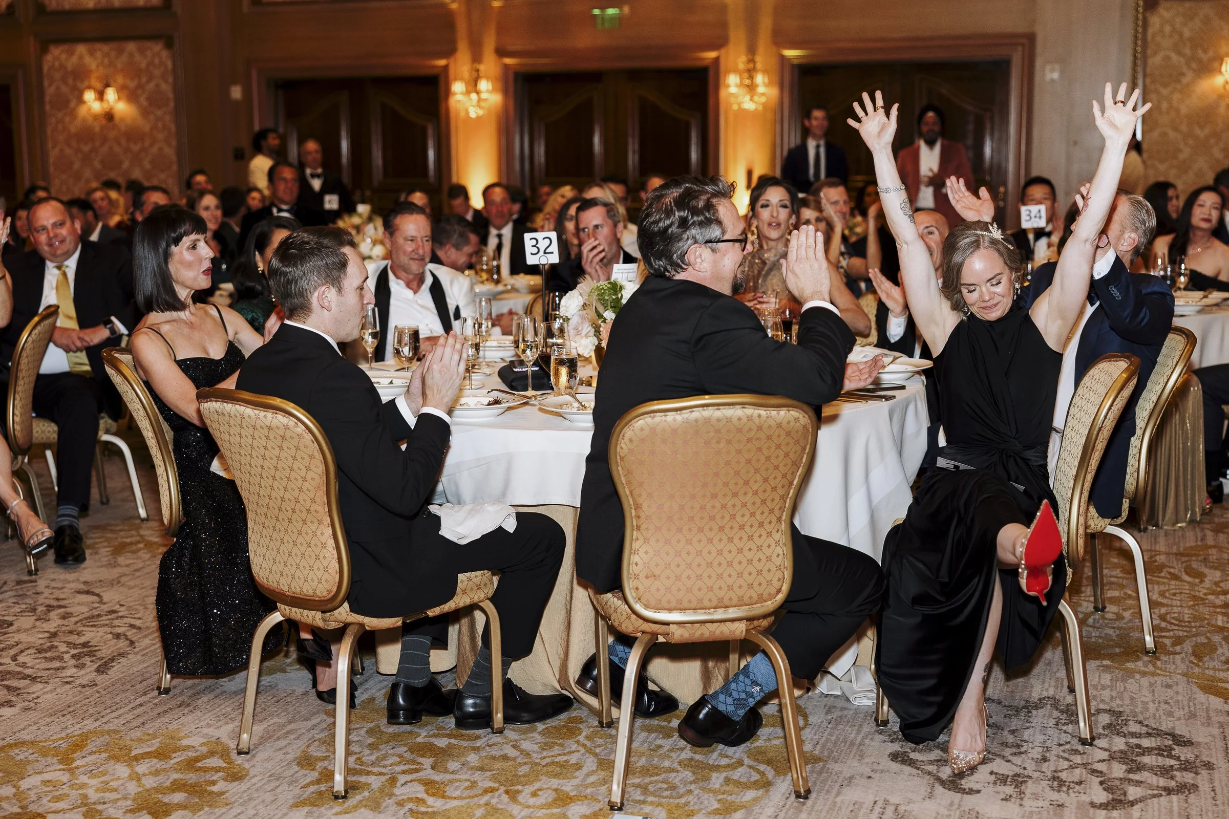 A group of formally dressed people sitting at round tables in a decorated banquet hall, with some raising hands and others smiling, during a celebration or event.