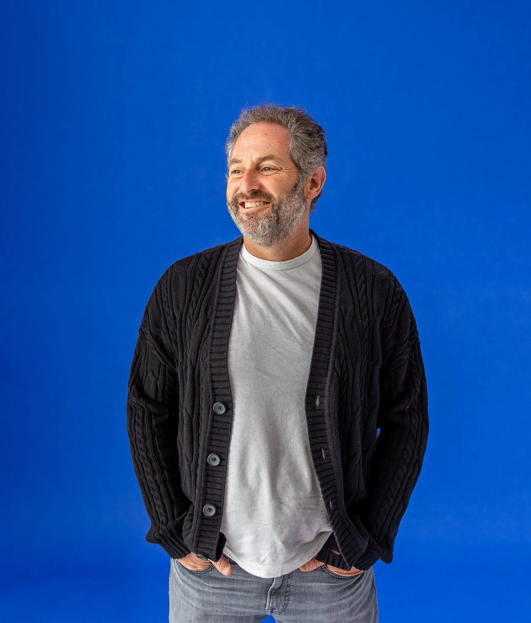 A man with gray hair and a beard, smiling, wearing a white t-shirt and a black knitted cardigan, standing against a solid blue background.