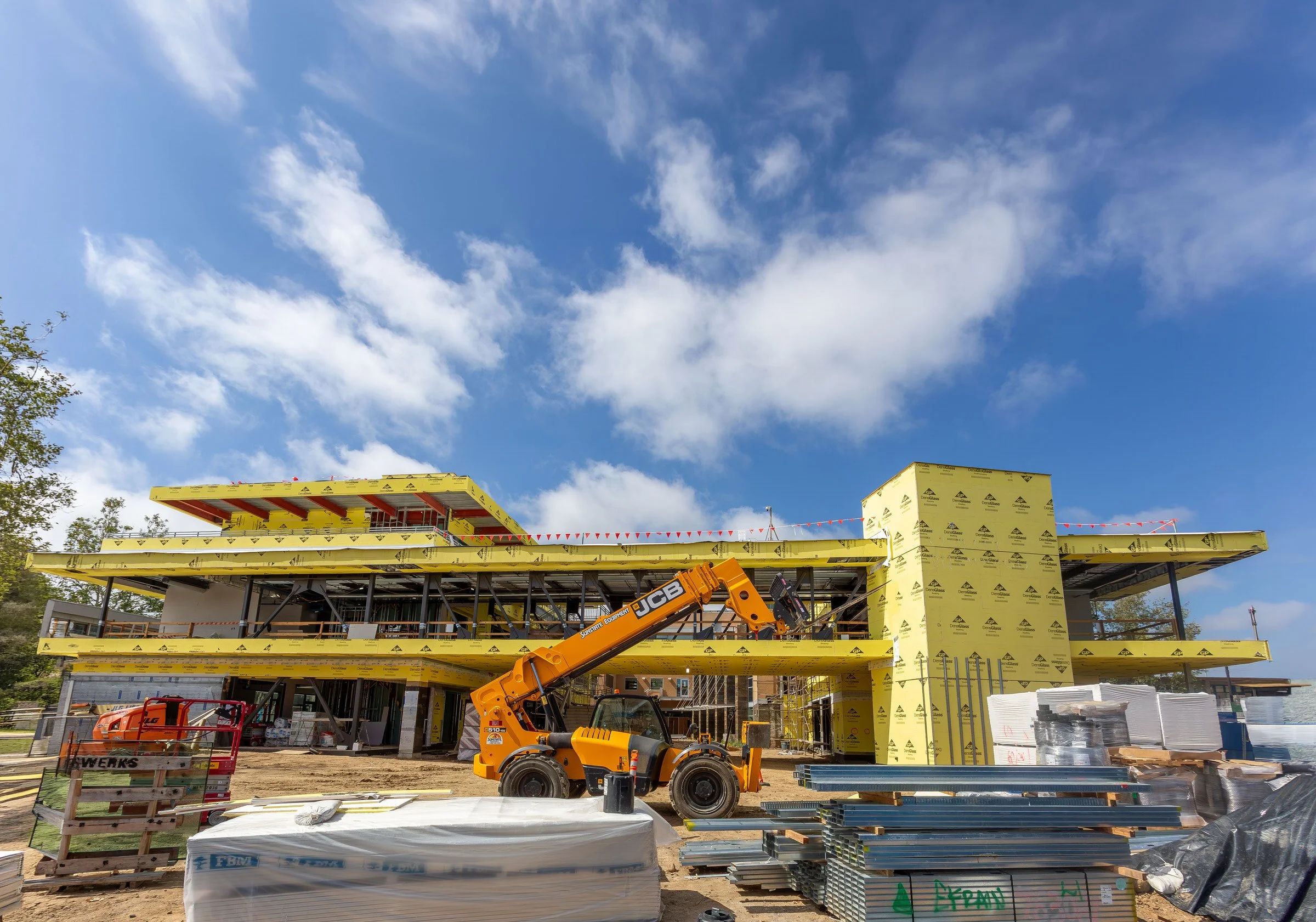 Construction site with a multi-story building under construction, scaffolding, a crane, and construction materials, with a bright blue sky with clouds overhead.