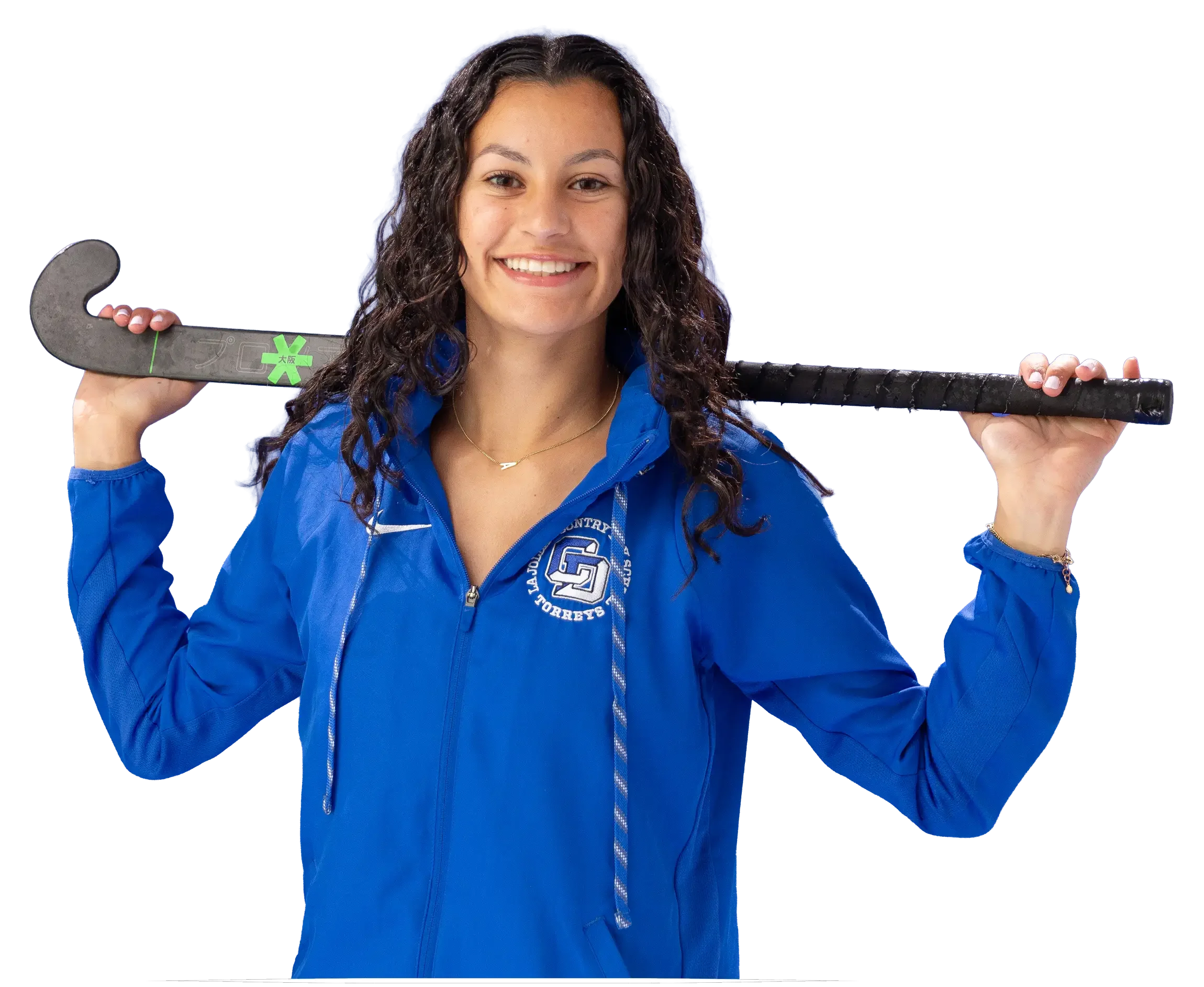 Young woman smiling, holding a black hockey stick over her shoulders, wearing a blue jacket with a logo, curly hair, and a necklace with a letter 'A' pendant.
