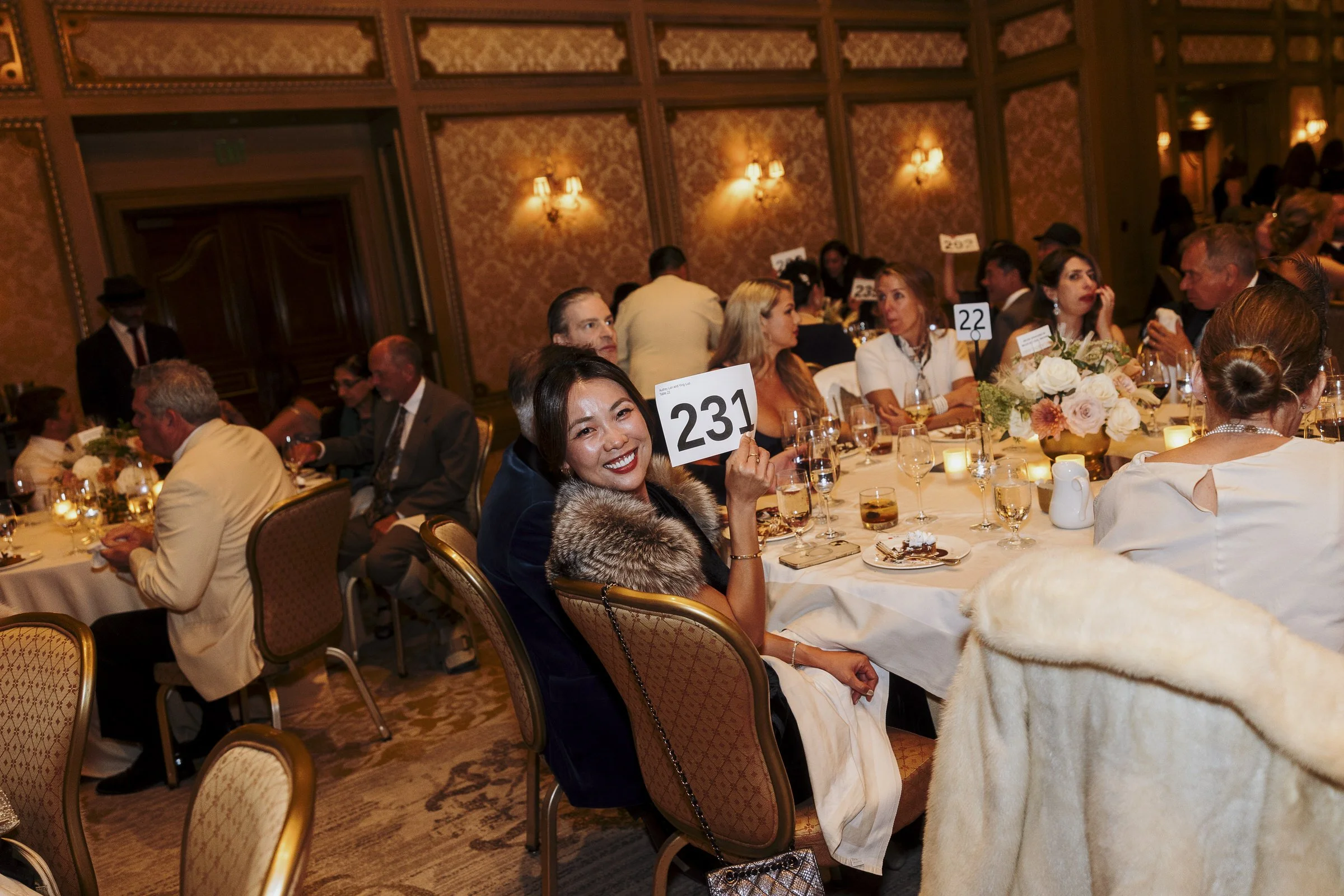 People sitting at round banquet tables during a formal event in an elegant room with ornate wallpaper and wall sconces, some holding up signs with numbers.