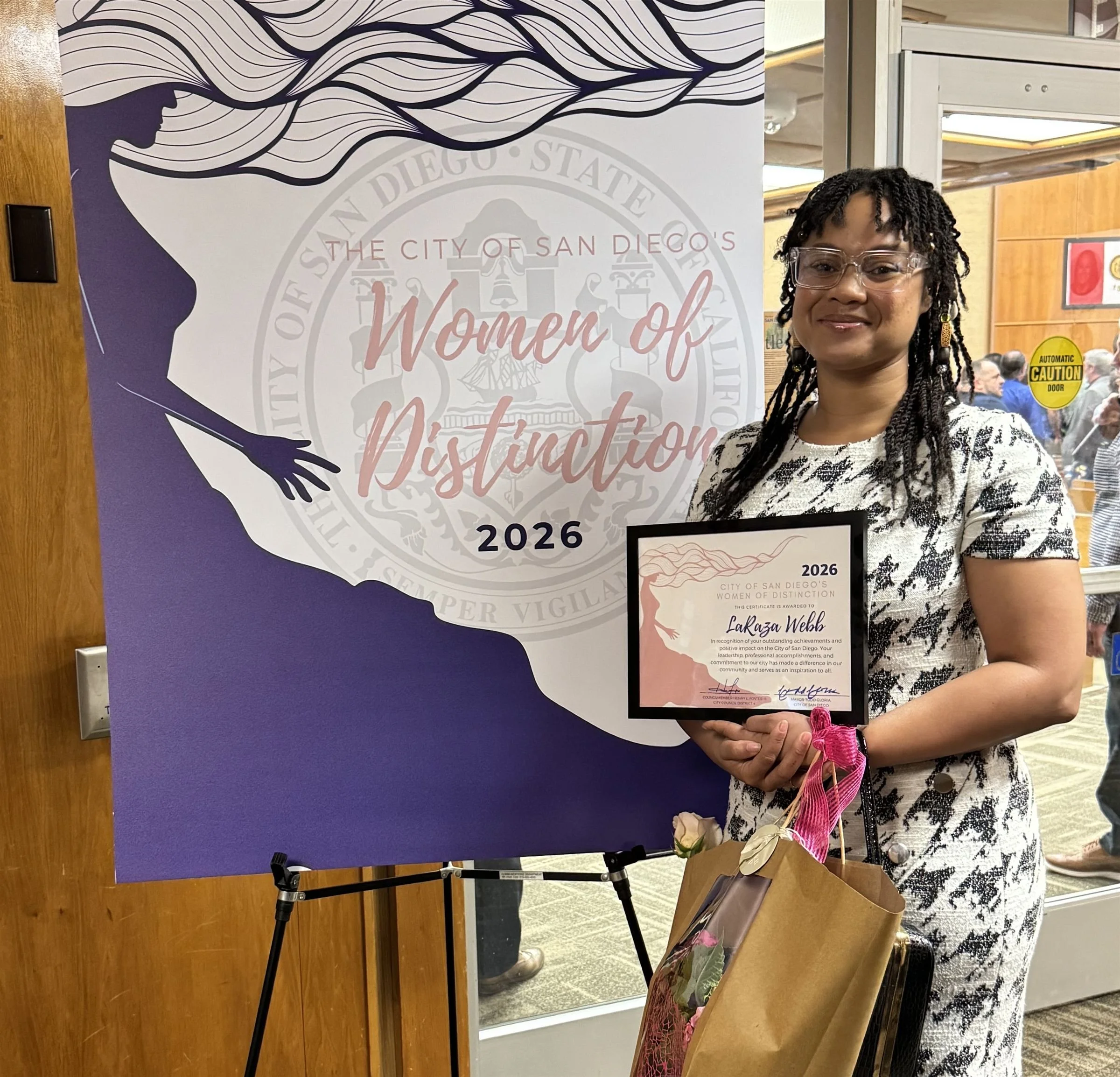 Woman holding a certificate standing next to a large banner for San Diego Women of Distinction 2026 event.