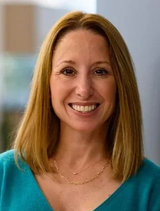 A woman with shoulder-length blonde hair smiling, wearing a teal top, indoors with a window in the background.