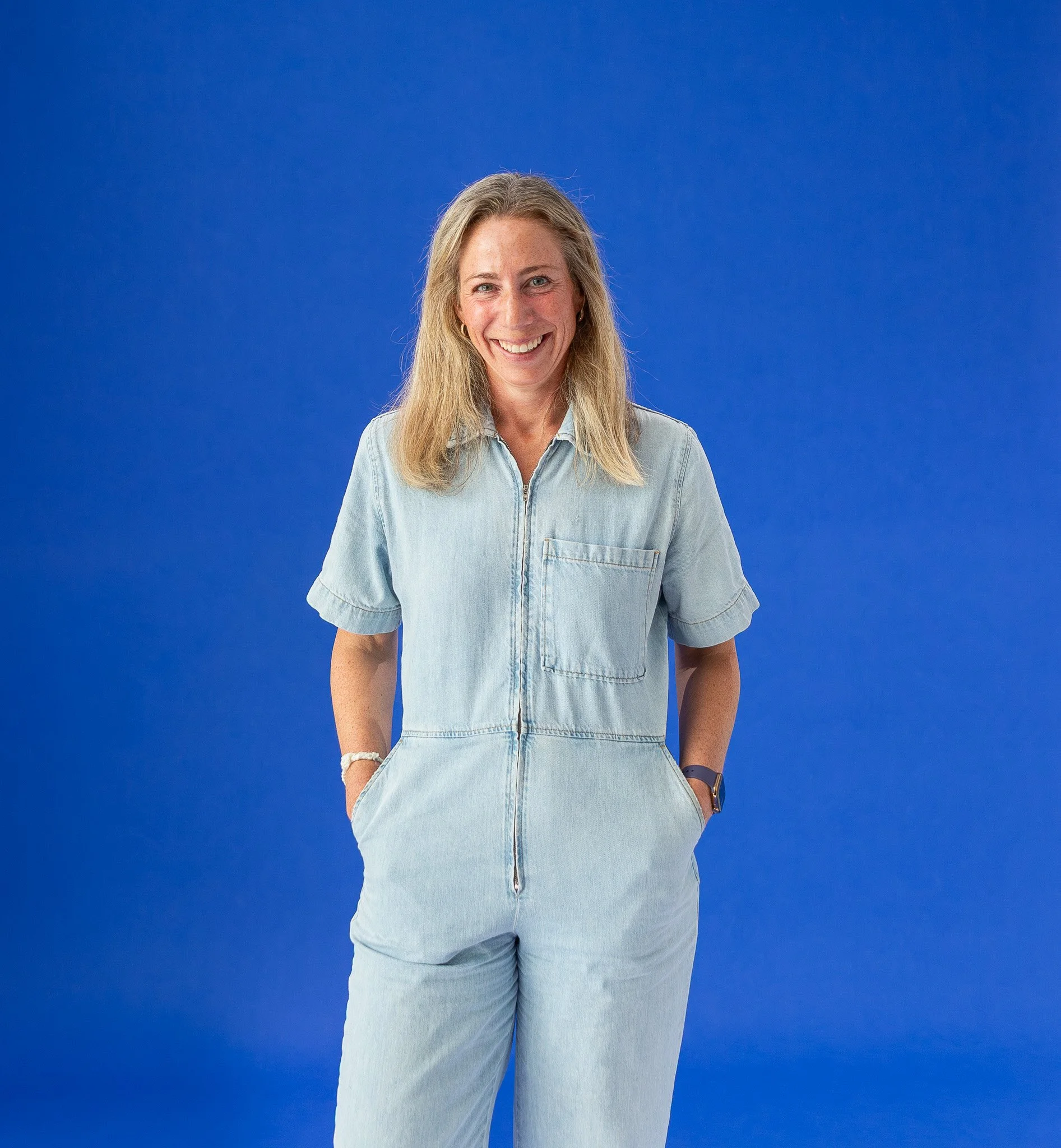 A woman with blonde hair smiling and wearing a light denim shirt with her hands in the pockets, set against a solid blue background.