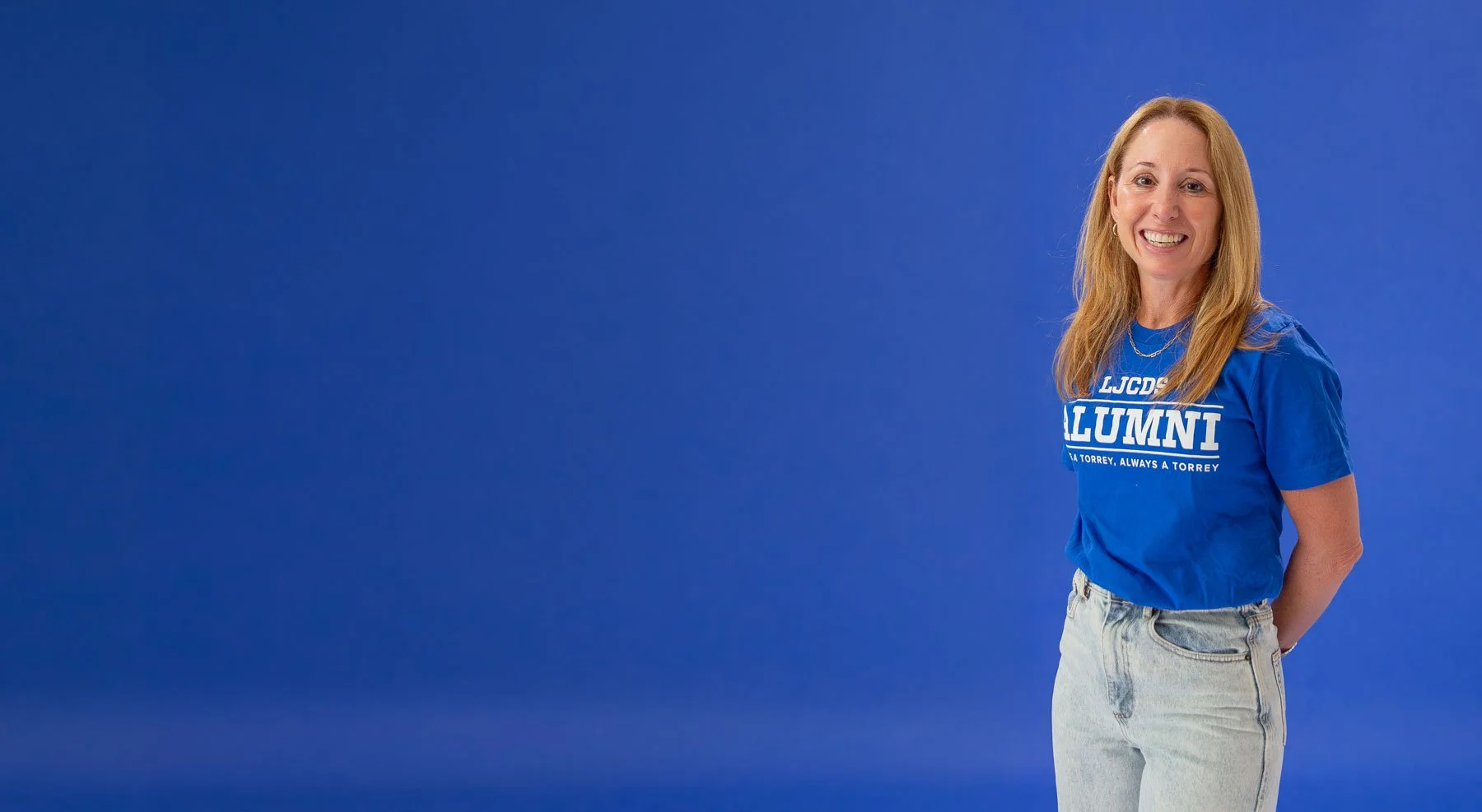 A woman with red hair smiling and standing with her hands behind her back, wearing a blue T-shirt with white text and light blue jeans, against a plain blue background.