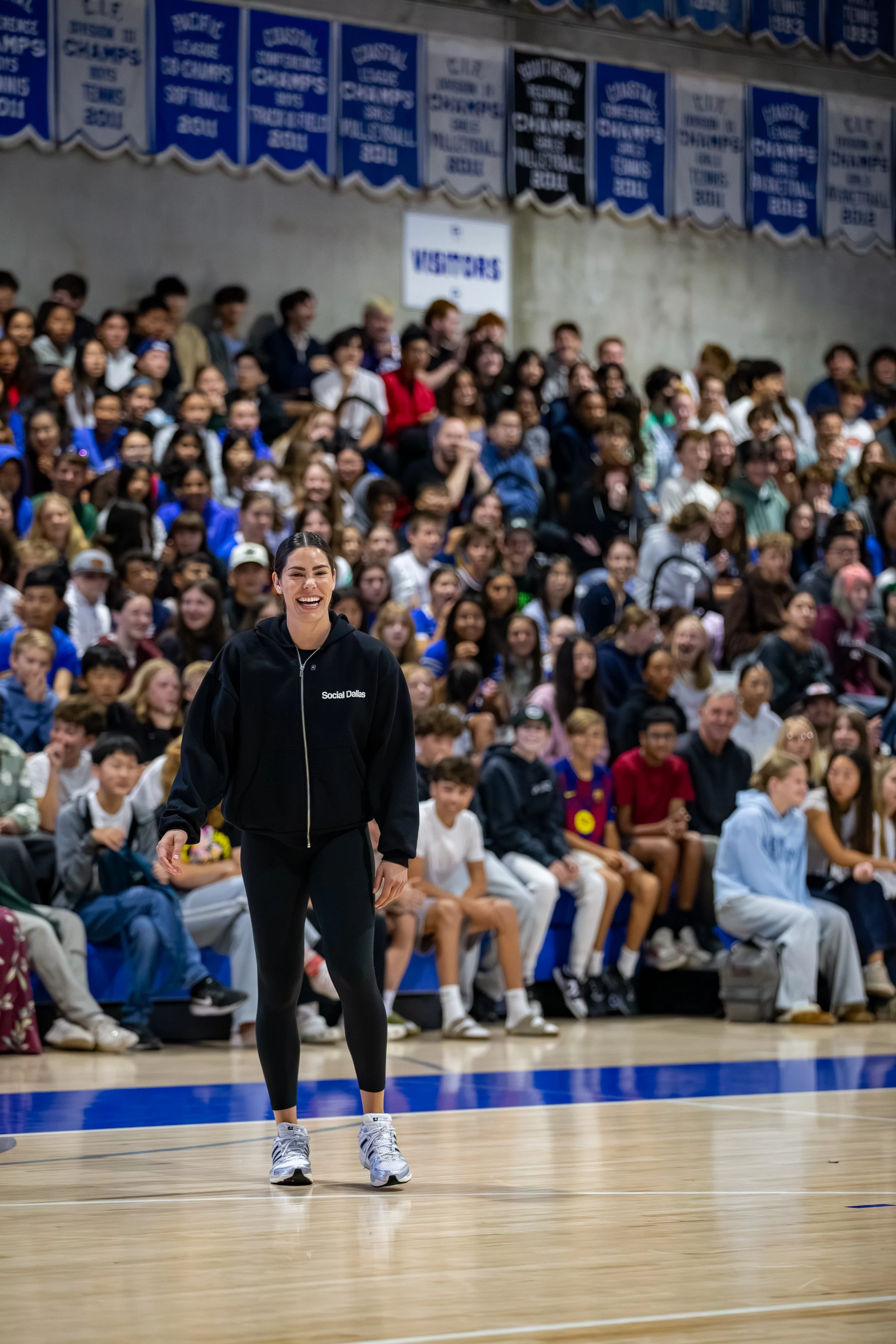 A woman in athletic wear standing on a gymnasium basketball court, smiling, with a large group of spectators seated behind her and banners hanging on the wall above.