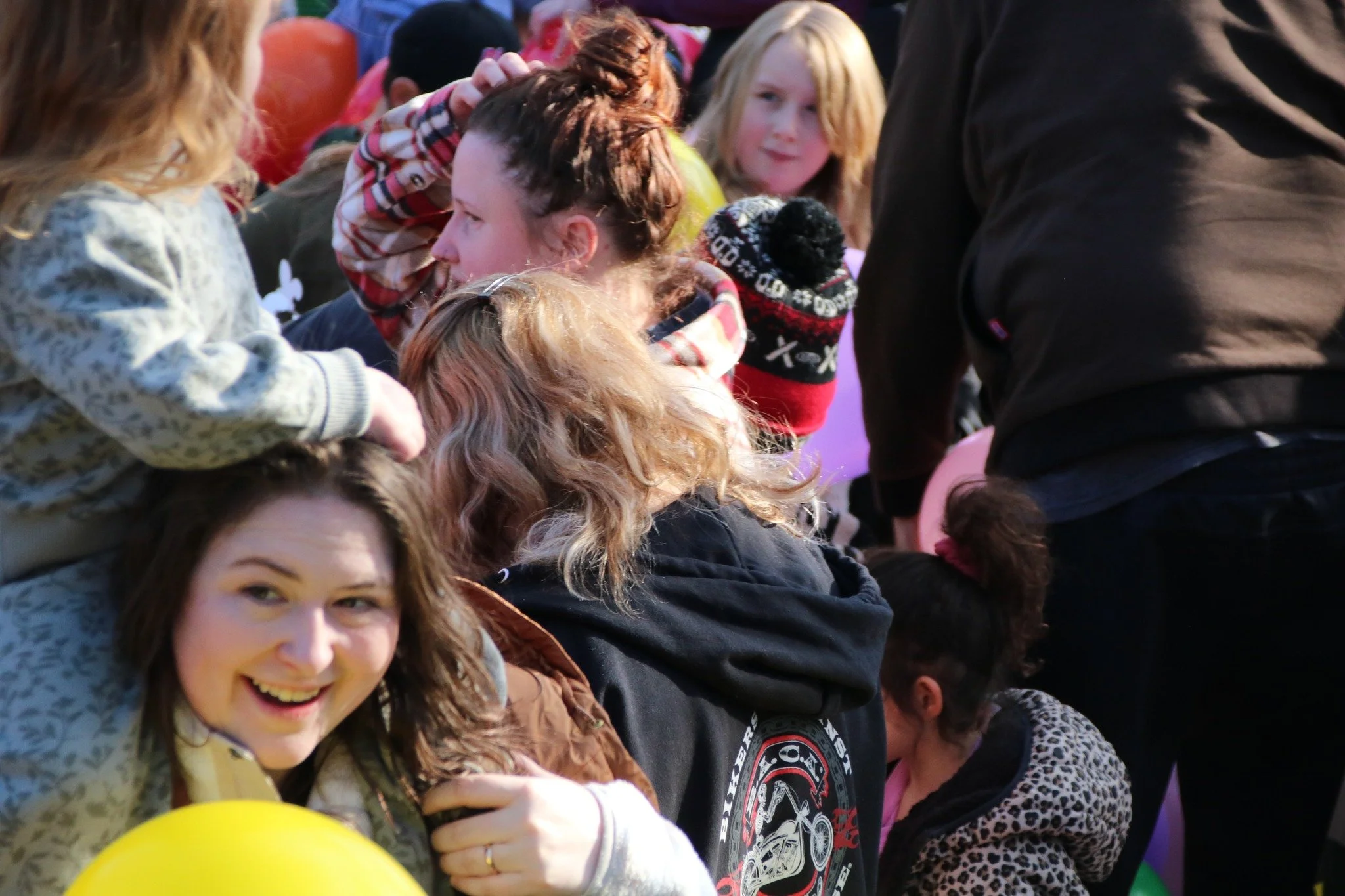Group of children and young adults at a crowded outdoor event, some with colorful balloons and winter clothing, interacting and smiling.
