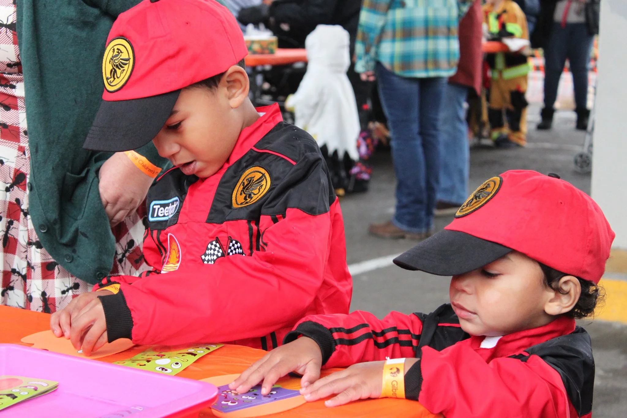 Two young boys dressed in racing jackets and red hats are sitting at a table, engaged in a craft activity. There are various colorful cards with cartoon faces on the table, and a pink tray in the foreground. People are in the background at an outdoor event.