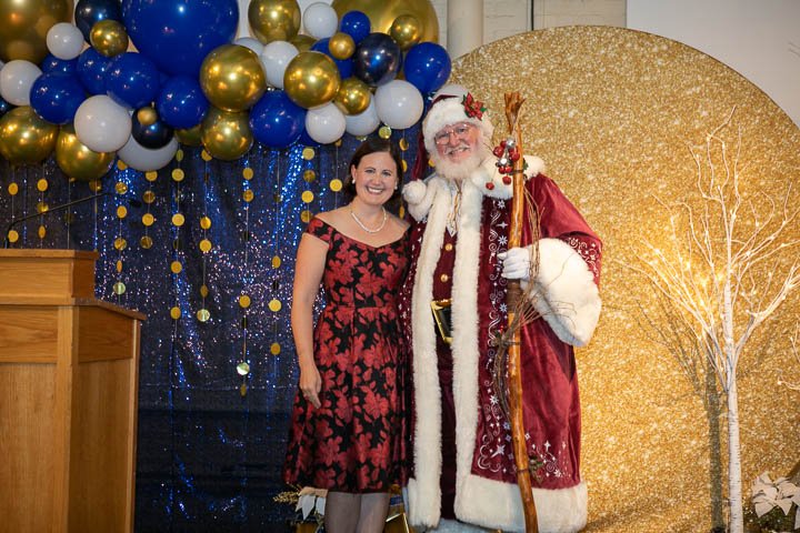 A woman in a red and black dress standing next to a man dressed as Santa Claus holding a staff, with a background of gold and blue balloons and a glittery, gold winter scene with a white tree.