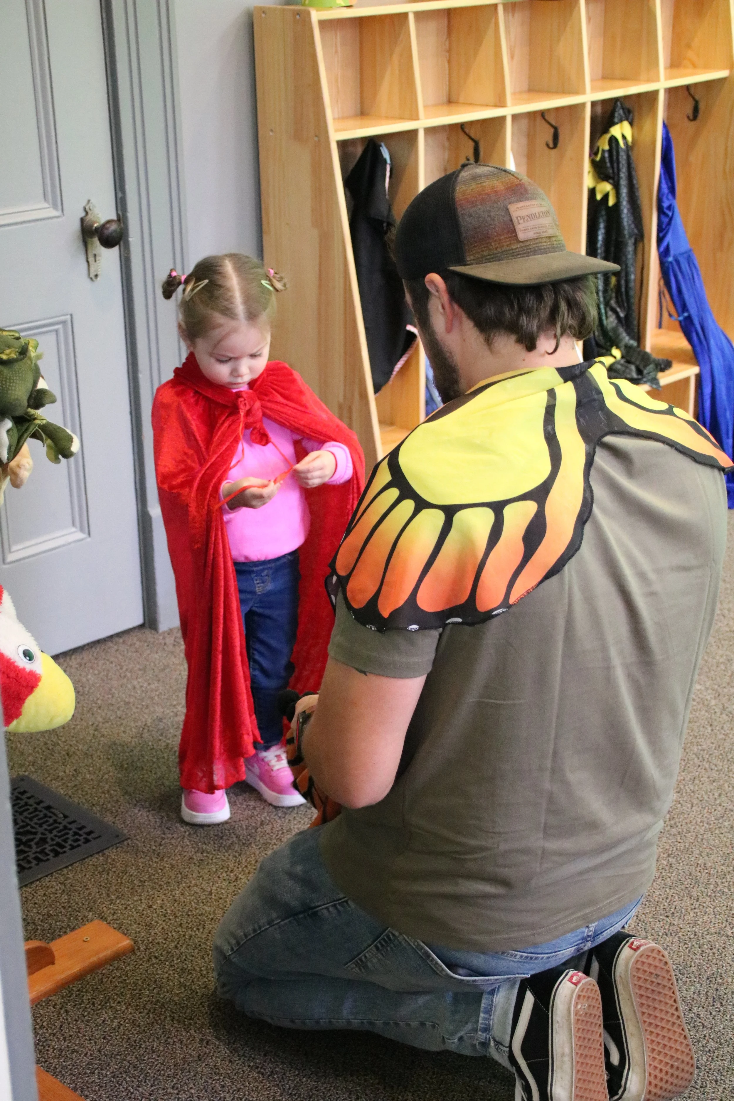 A man kneeling on the floor dressed in a costume with butterfly wings talking to a young girl wearing a red cape and school uniform. The girl is holding a small object and is inside a room with wooden cubbies and hooks in the background.