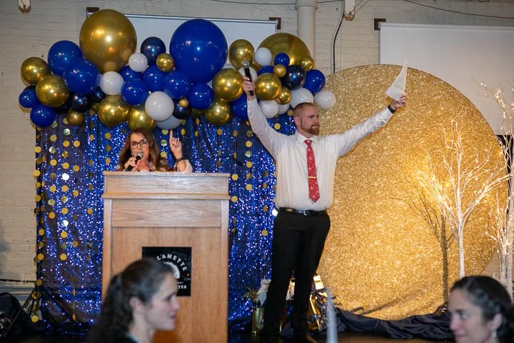 A woman speaking at a wooden podium with conducting microphone, and a man holding a microphone and pointing upward, standing on a decorated stage with gold and blue balloons, blue curtain backdrop with gold dots, and a large gold glitter circle with branches in the background.