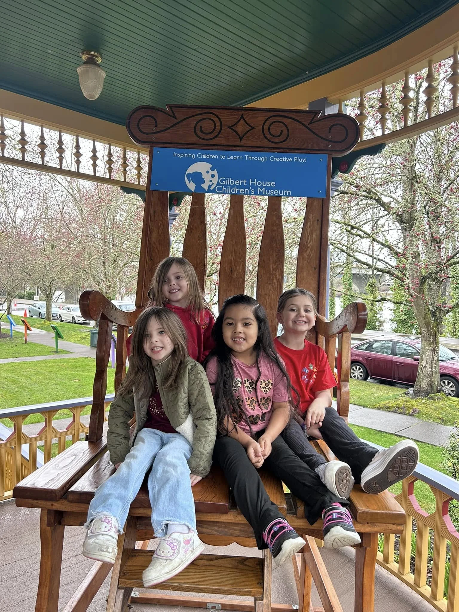 Four young girls sitting on a giant wooden chair at the Gilbert House Children's Museum, with trees and parked cars in the background.