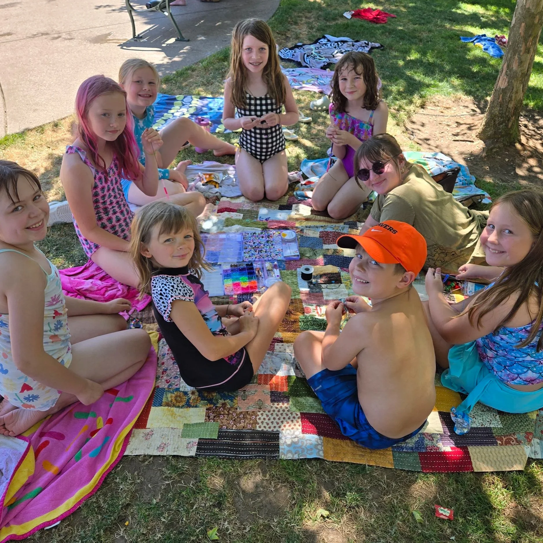 Group of children sitting on a patchwork quilt outdoors under a tree, enjoying a sunny day, with towels and clothes scattered around.