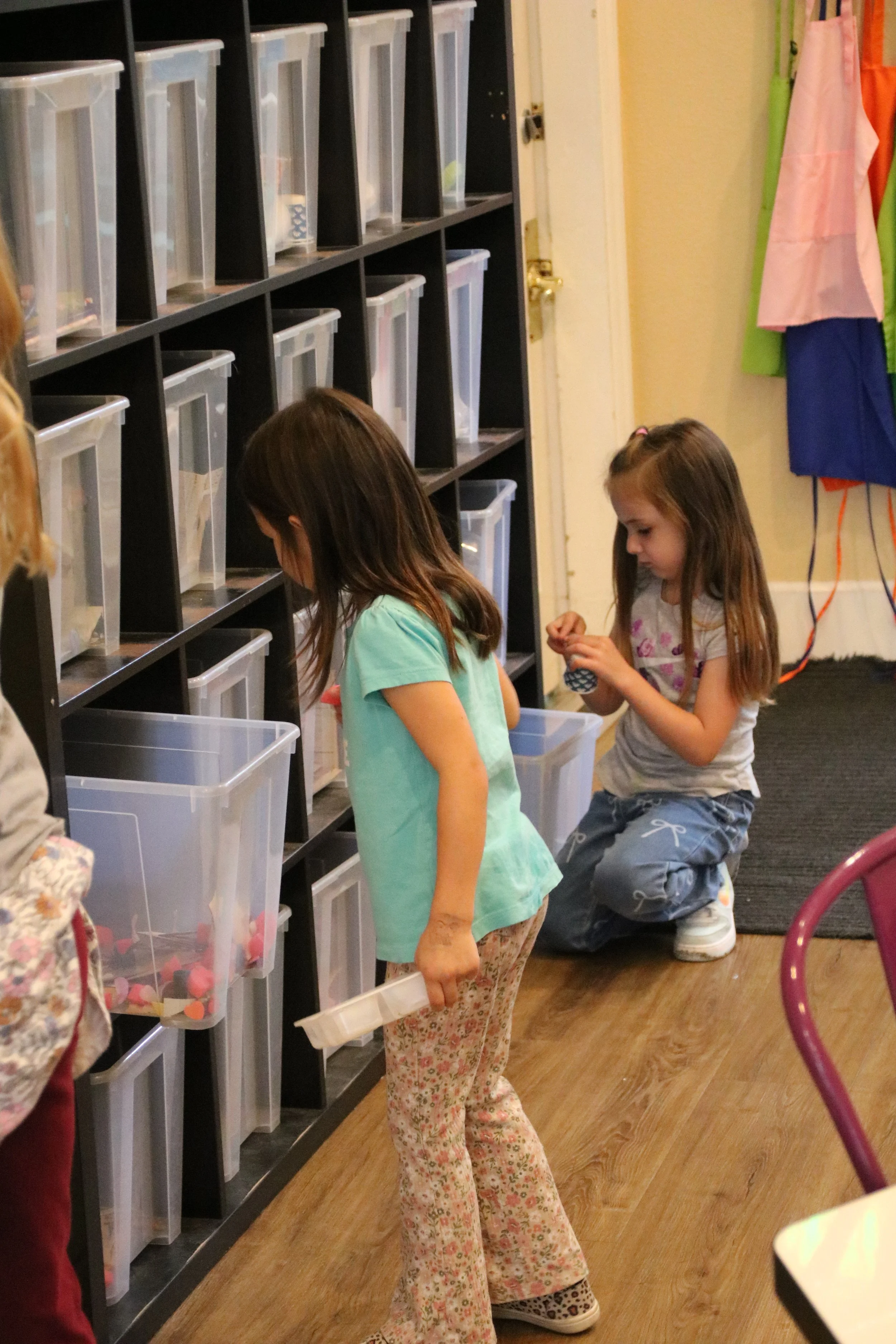Two young girls are at a sorting station with labeled plastic bins on a black shelf. One girl is holding a clear container, while the other girl is kneeling and picking items from a bin.