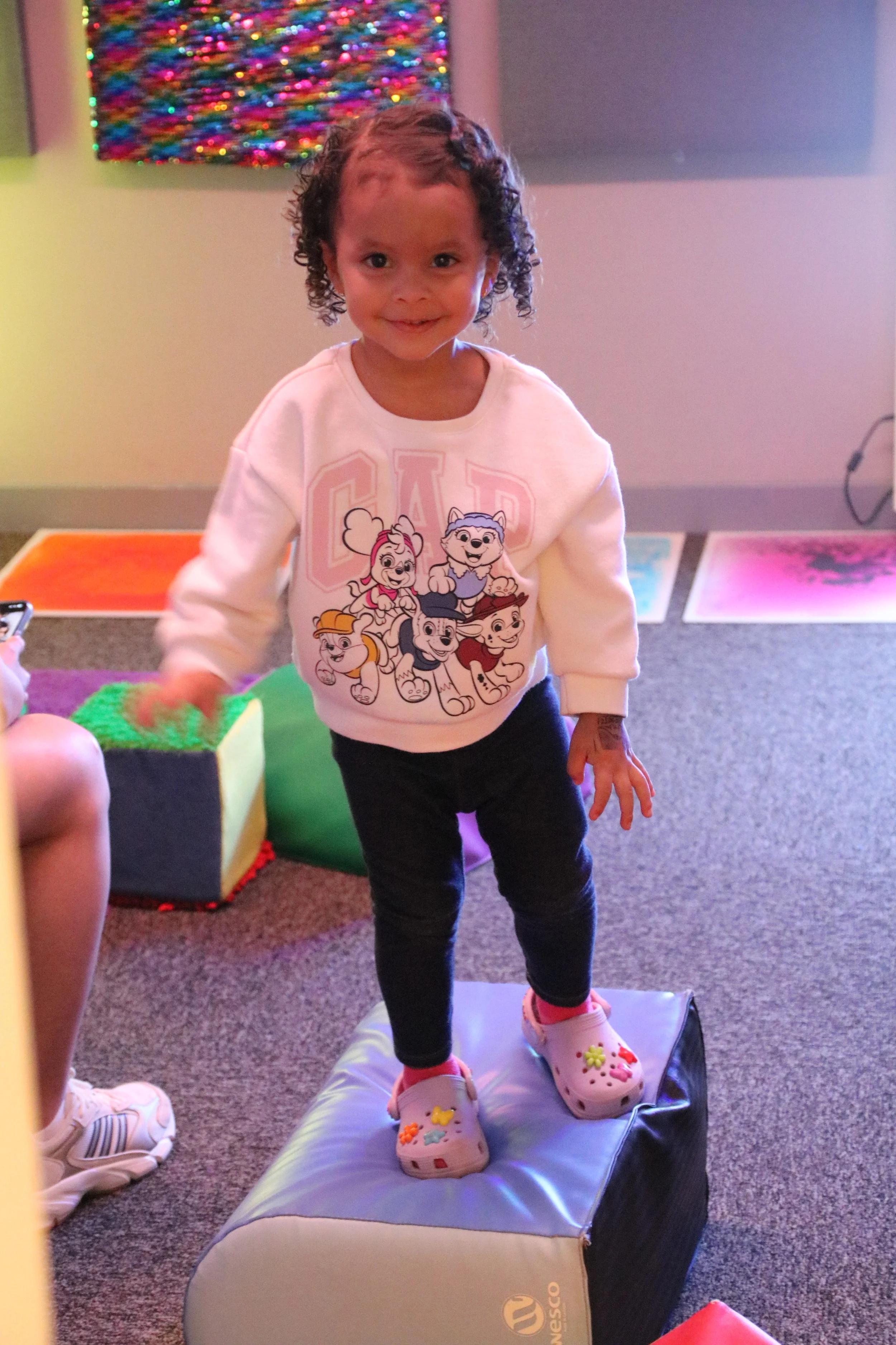 A young girl with curly hair smiling, standing on a padded stool, wearing a white sweatshirt featuring cartoon characters, black pants, and pink Crocs decorated with colorful charms, in a room with colorful wall art.